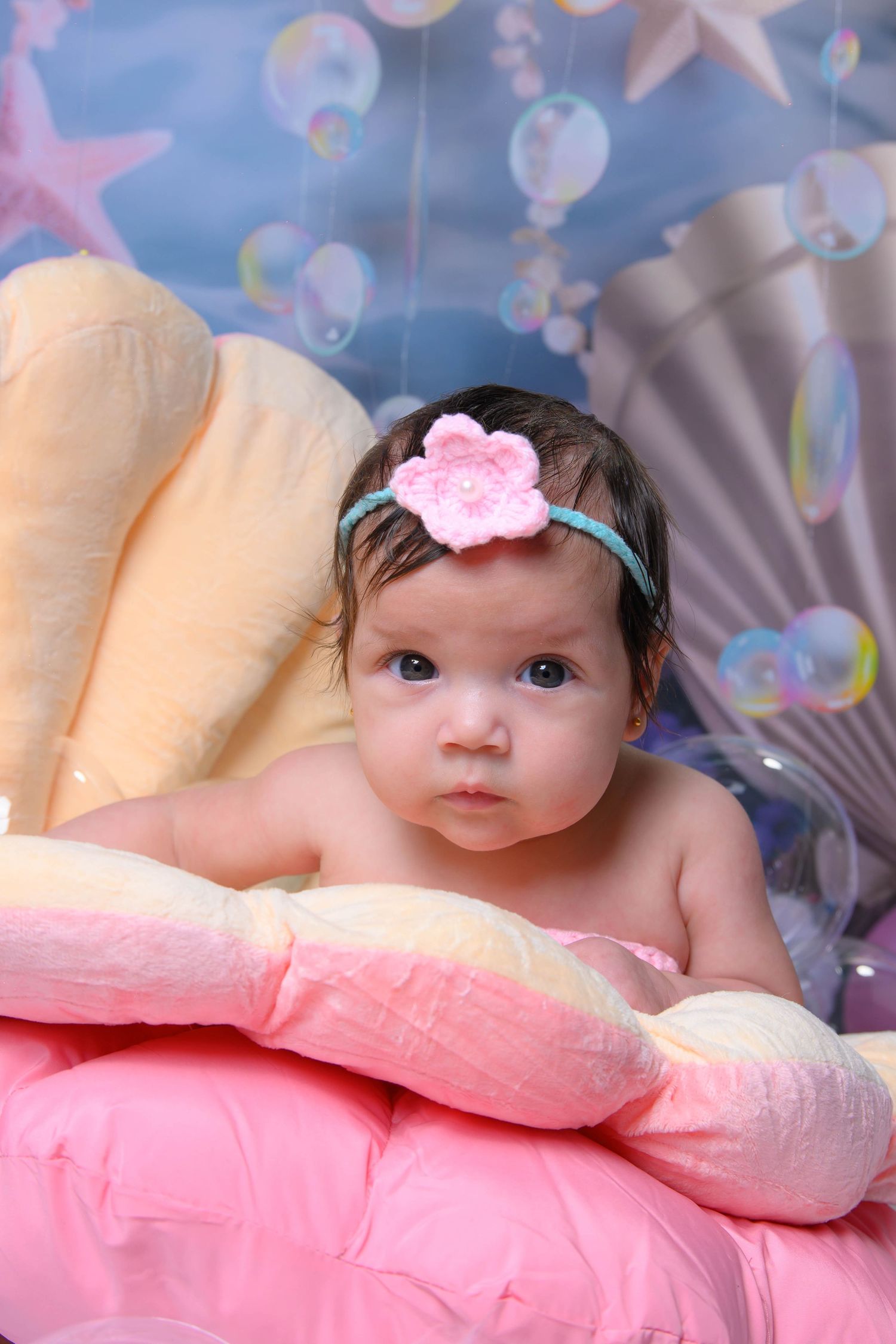 Baby in pink headband lying on soft pink blanket with dreamy blue starry backdrop.