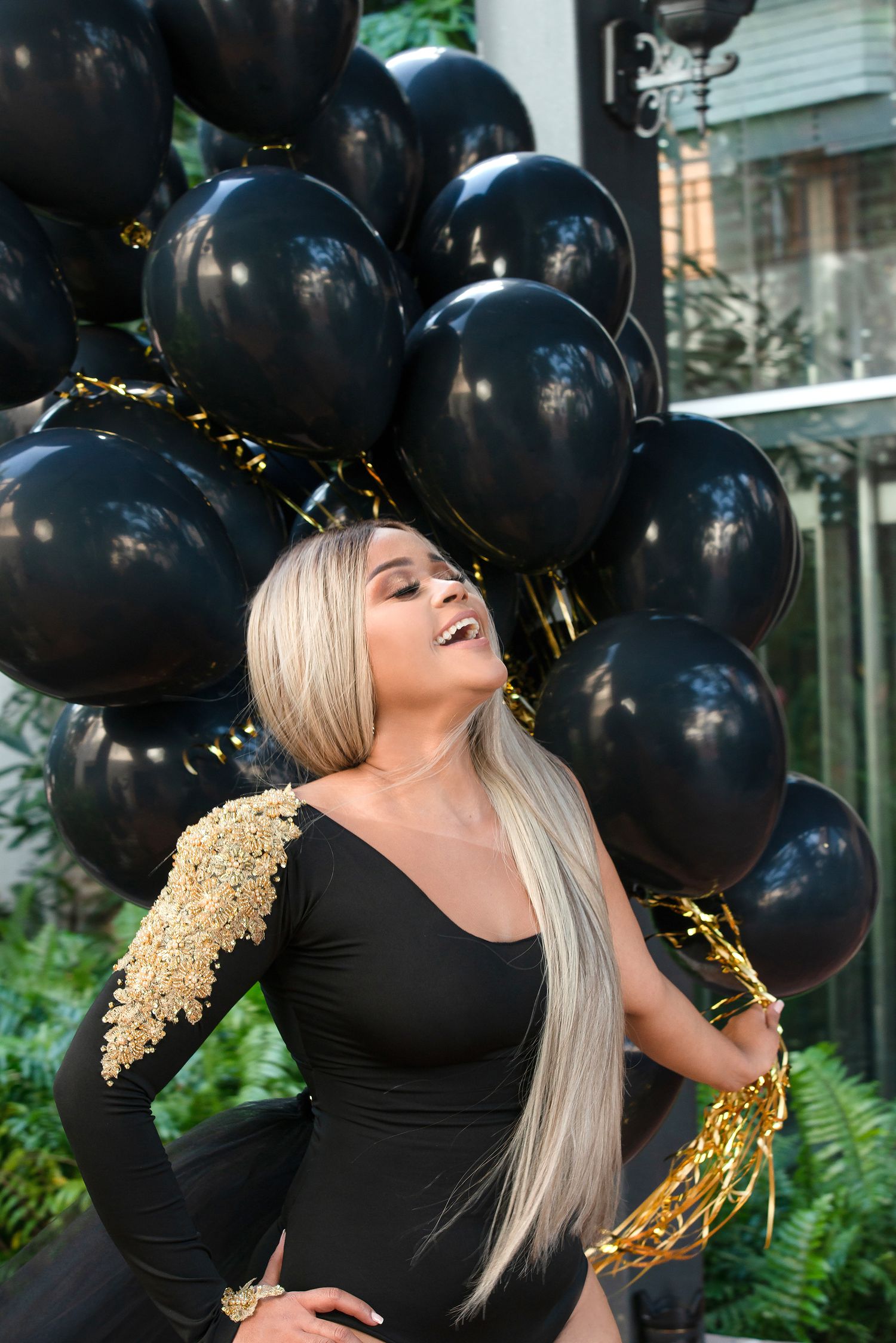 Woman in black dress laughs joyfully surrounded by black balloons and gold confetti against green foliage backdrop.