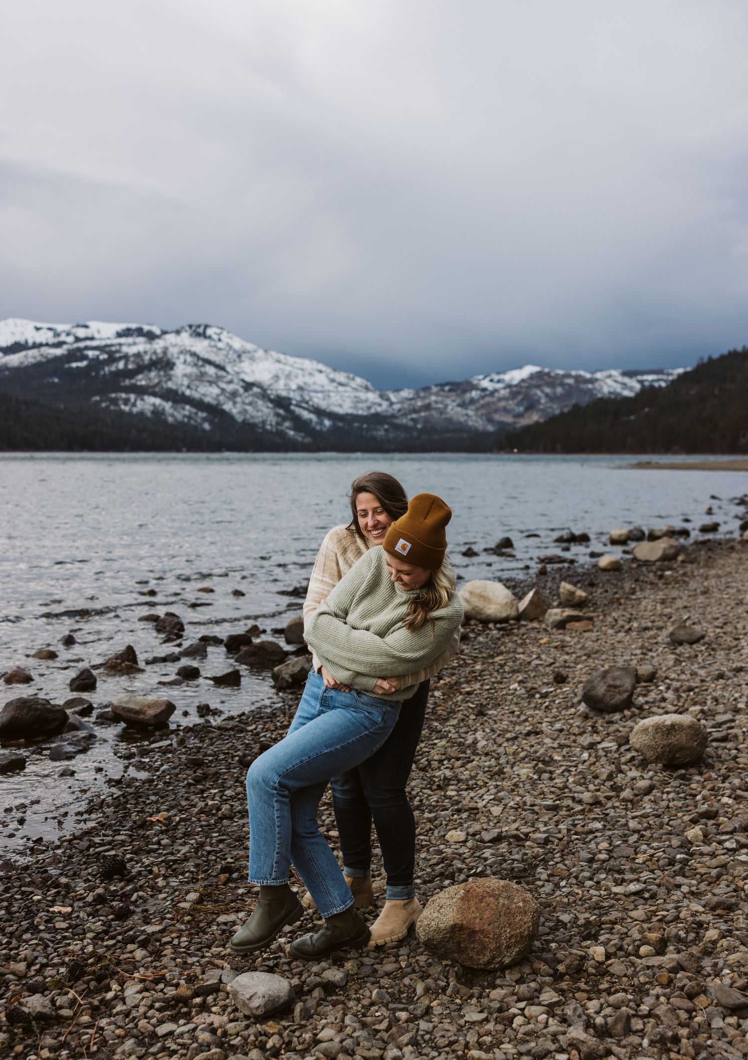 Couple sitting on rocky beach shore with snow-capped mountains and cloudy skies in the background.