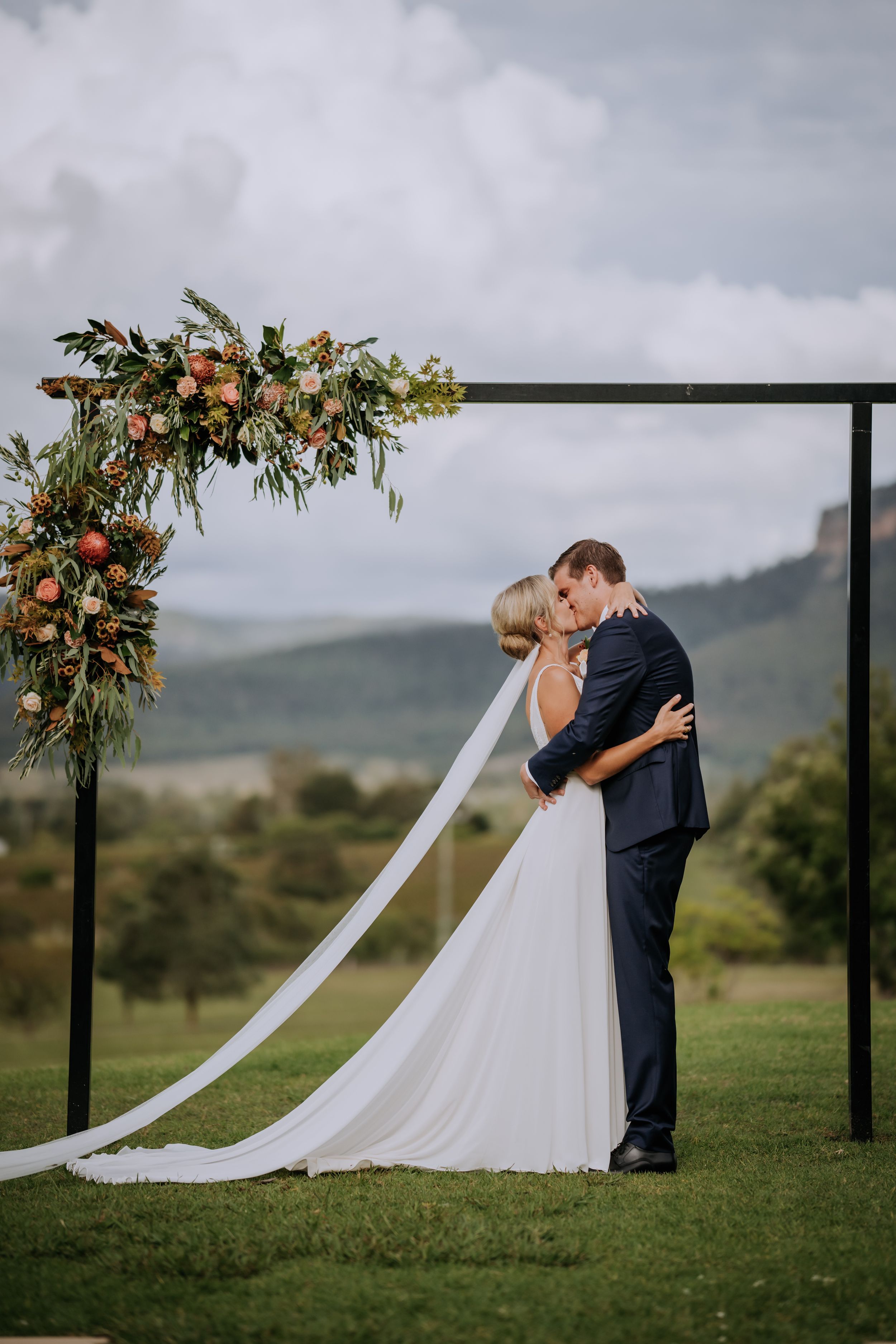 Wedding couple sharing intimate moment on hillside with flowing white dress and floral arch.
