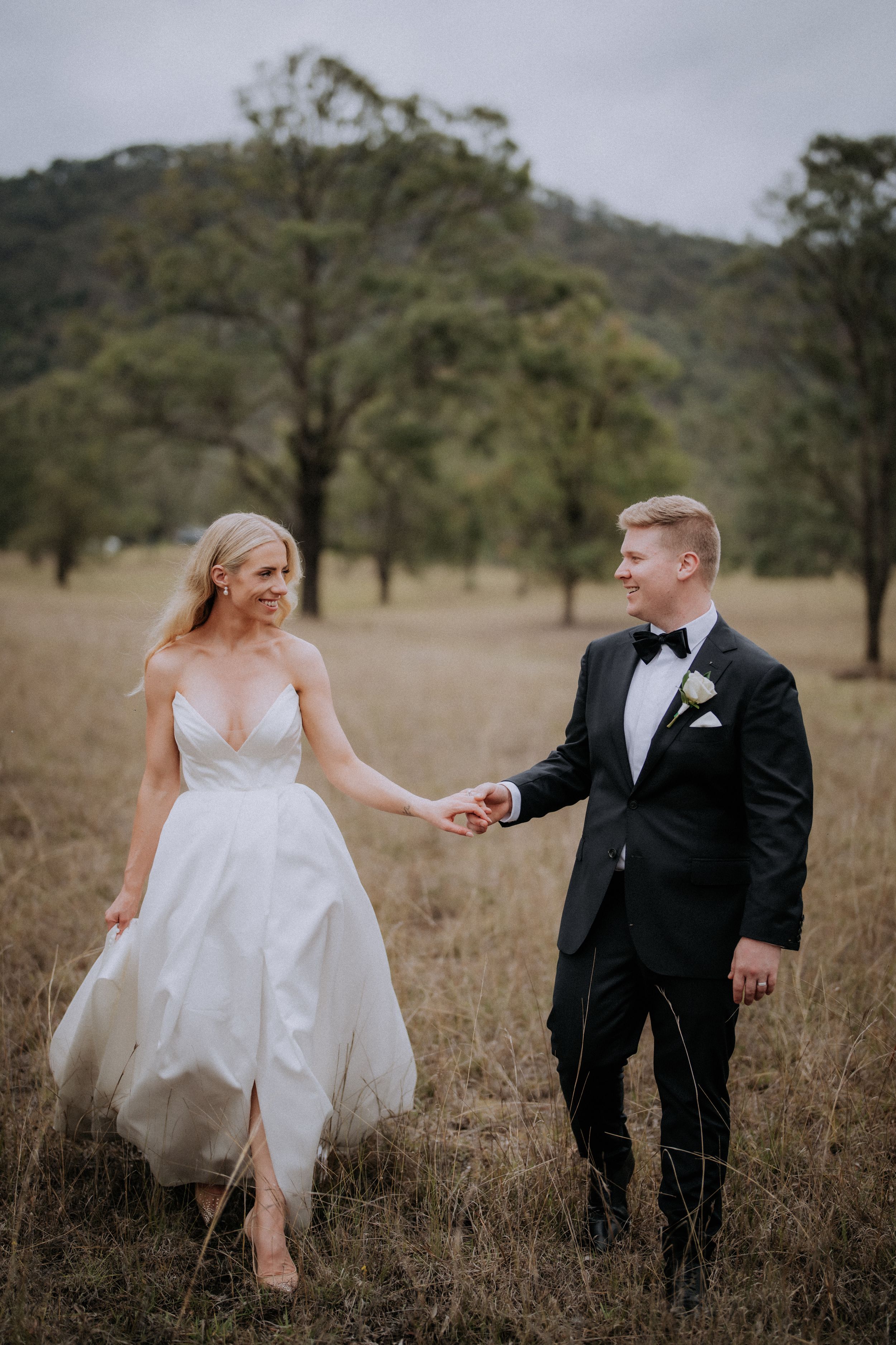 Newlyweds walk hand in hand through a rustic field with scattered trees in the background.