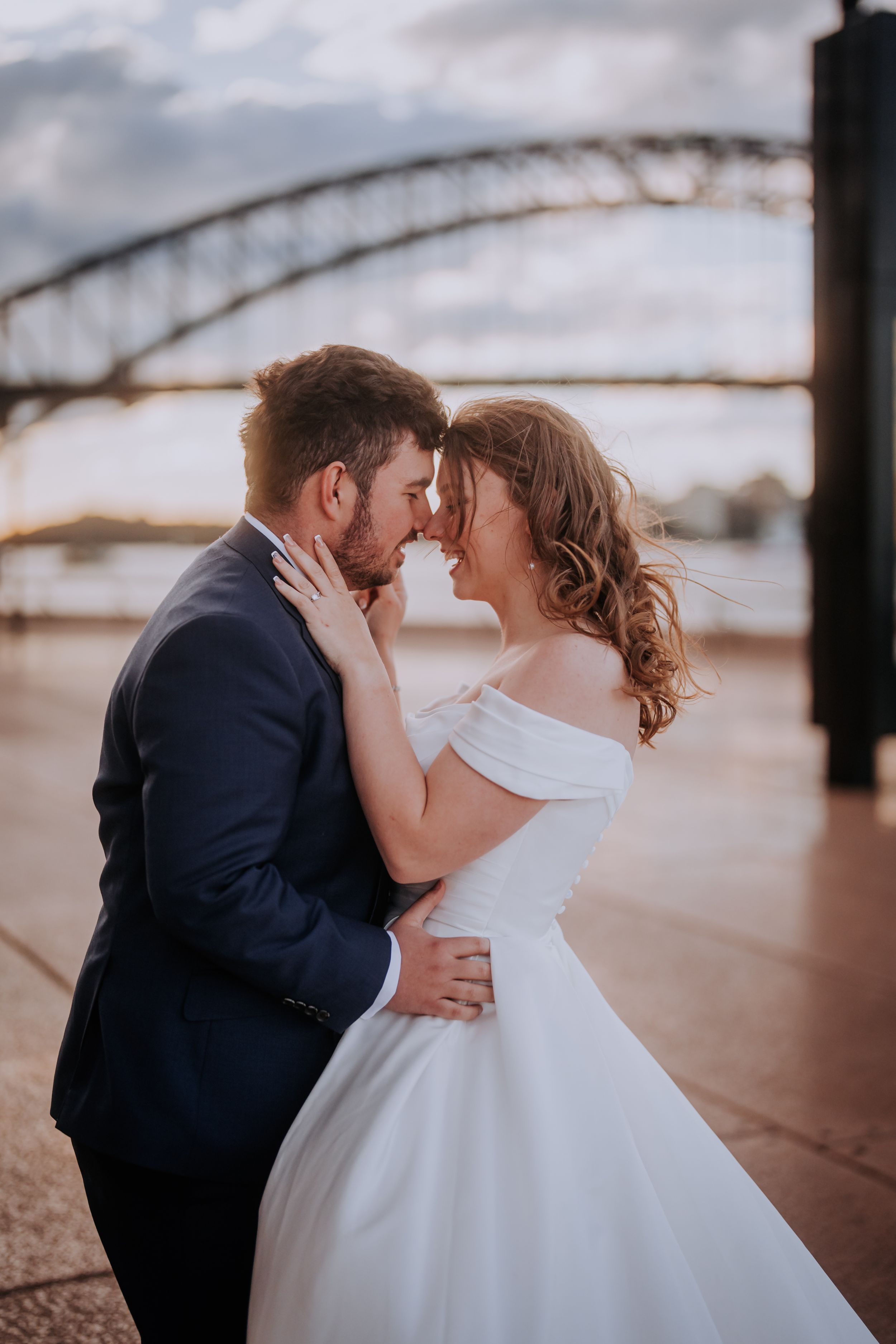 Romantic couple in wedding attire embrace near bridge during golden hour.