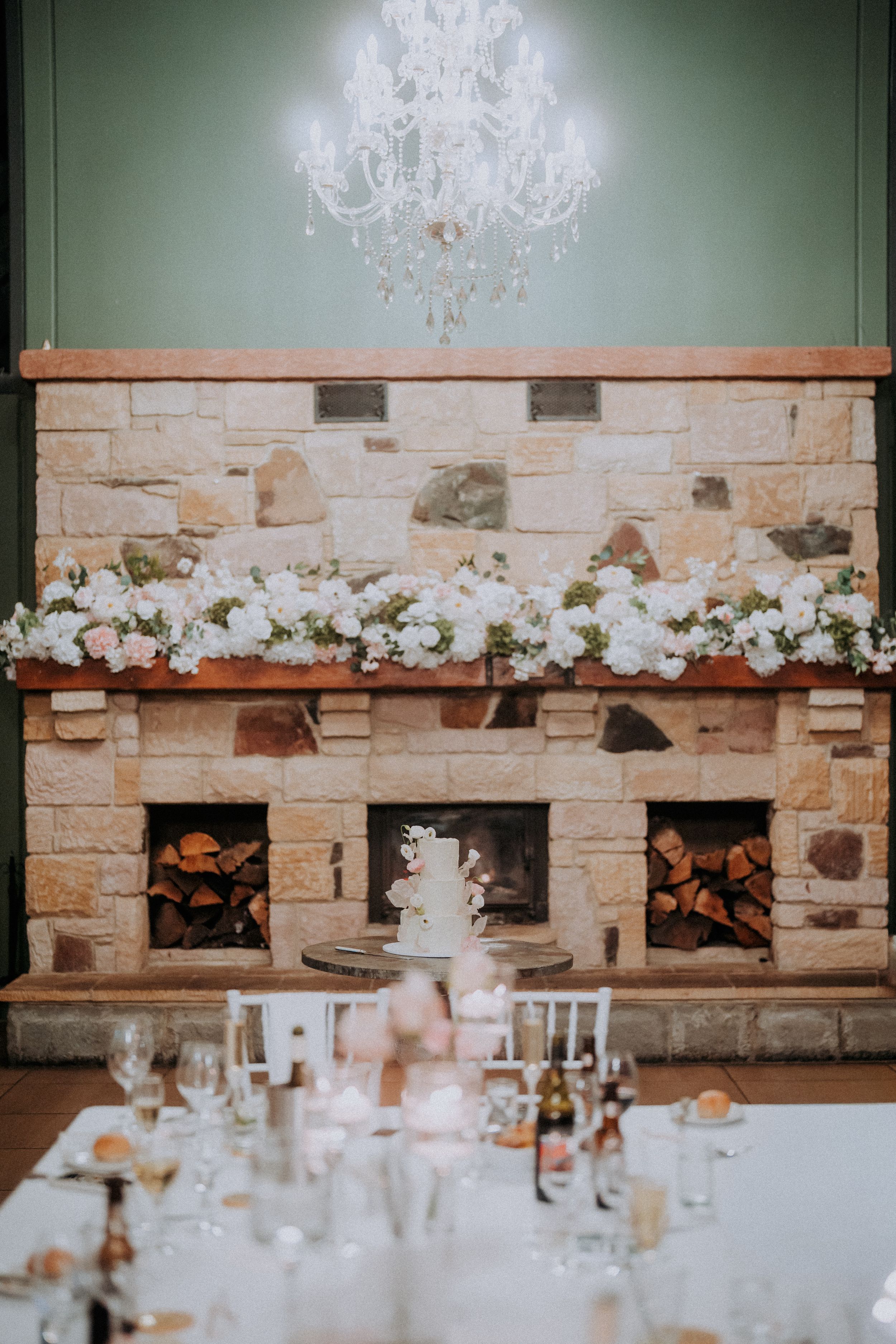 Elegant wedding reception decor with crystal chandelier and stone fireplace adorned with white flowers.