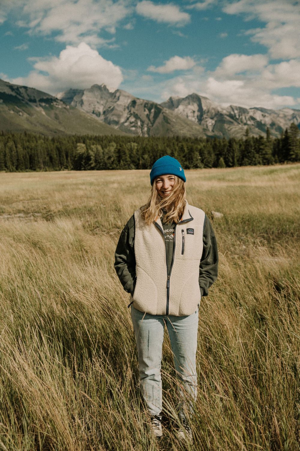 Person wearing a blue beanie and light colored vest stands in tall grass with mountains in the background.