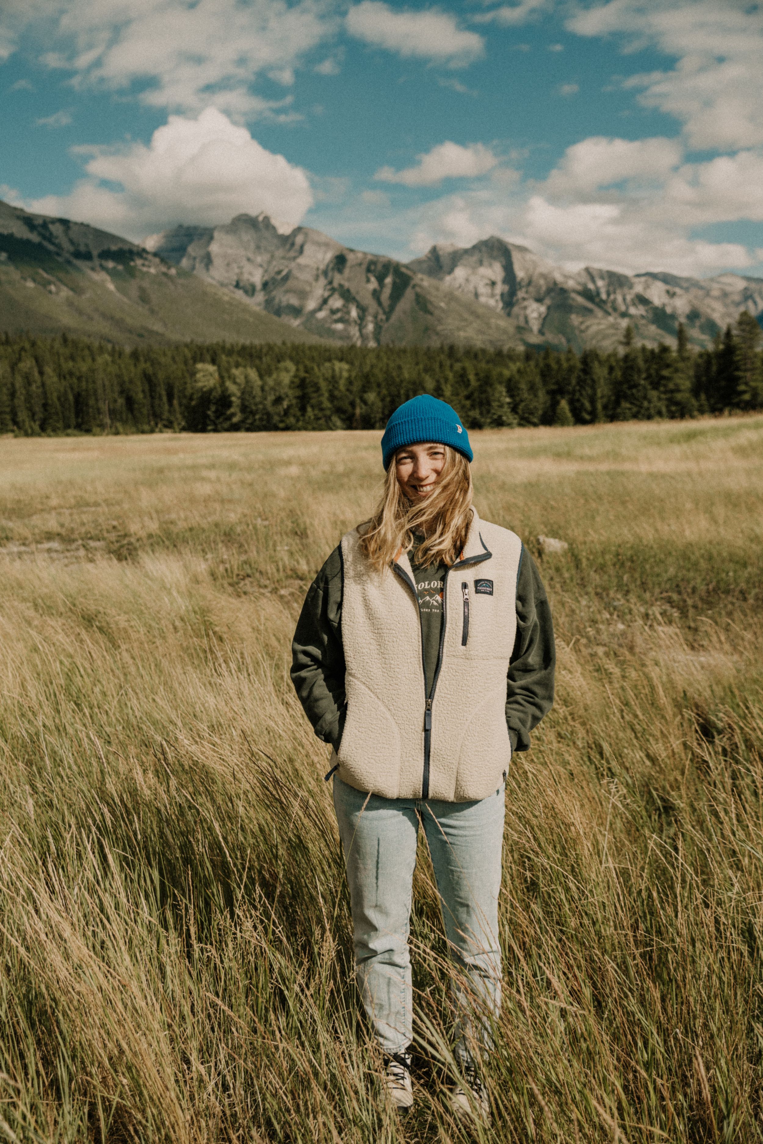 Person wearing a blue beanie and light colored vest stands in tall grass with mountains in the background.