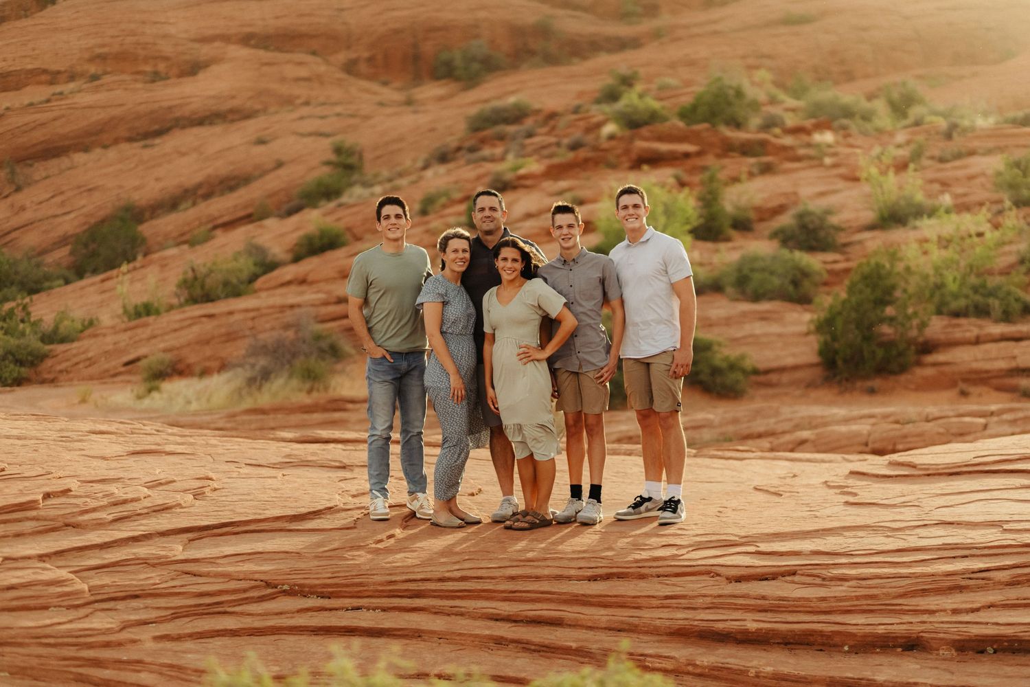 A group poses for photos on red sandstone rock formations in a desert landscape at sunset.