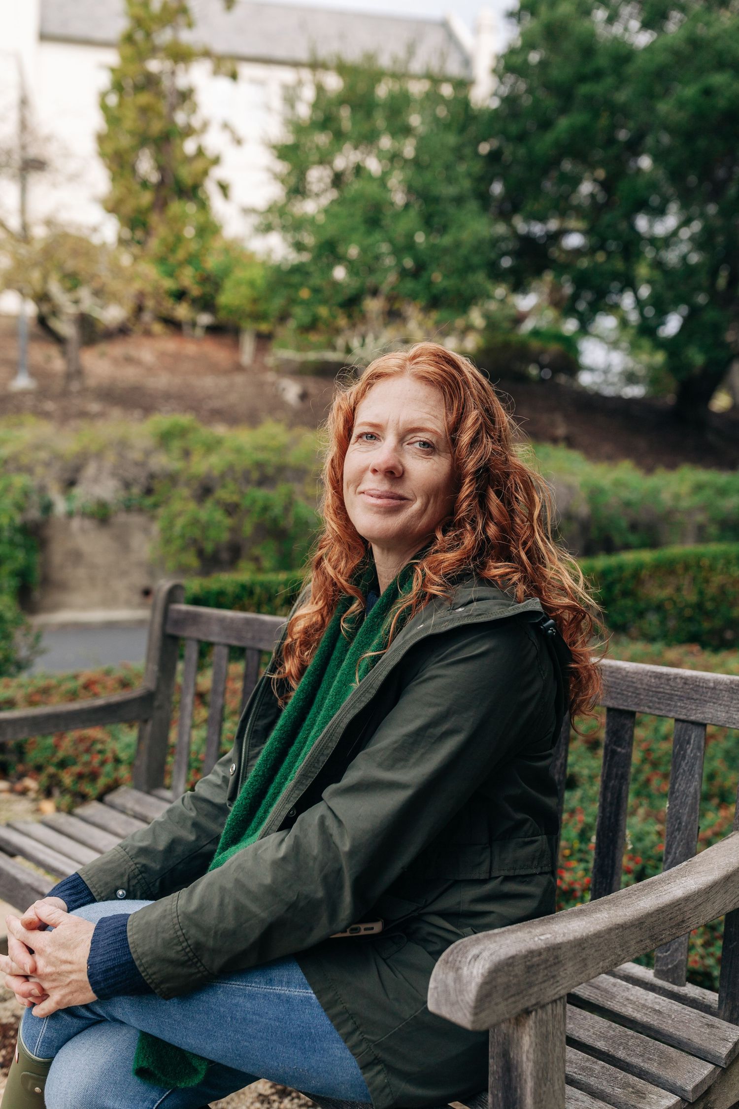 A casual outdoor portrait featuring someone in a green scarf and dark jacket sitting on a wooden bench.