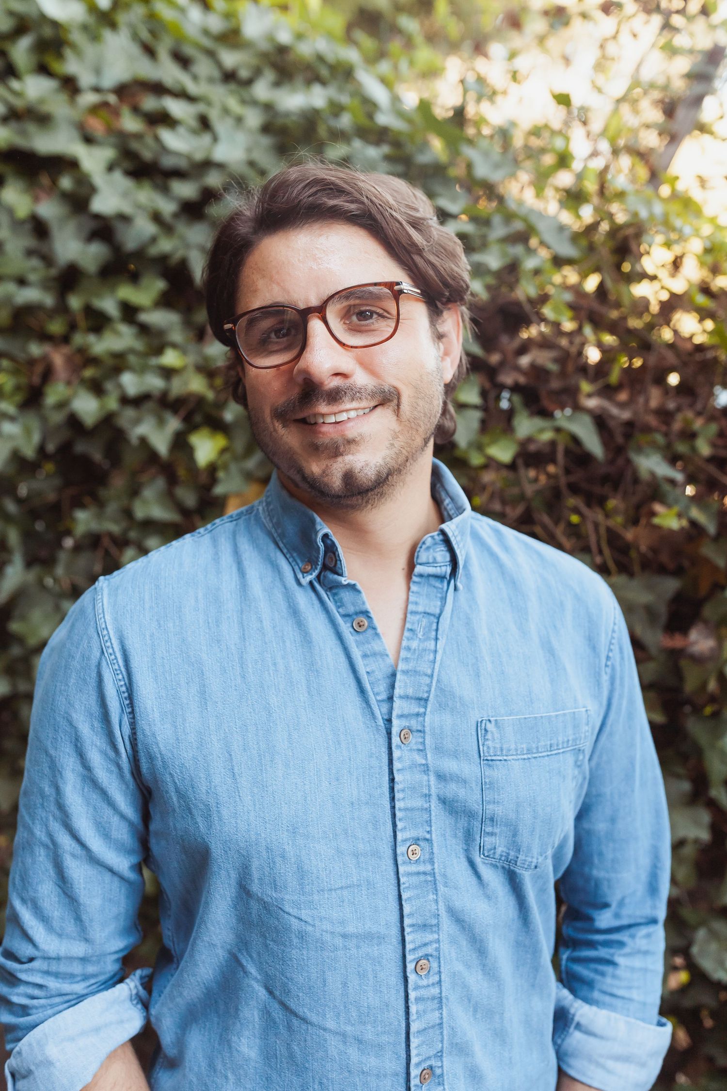 Casual portrait of someone in denim shirt and glasses against greenery.