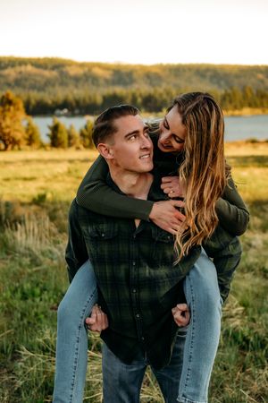 A playful piggyback ride moment between a couple in an autumn field by a lake.