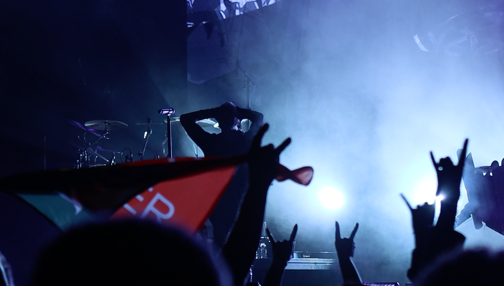 Concert crowd silhouettes raise their hands during an energetic performance with blue stage lighting.