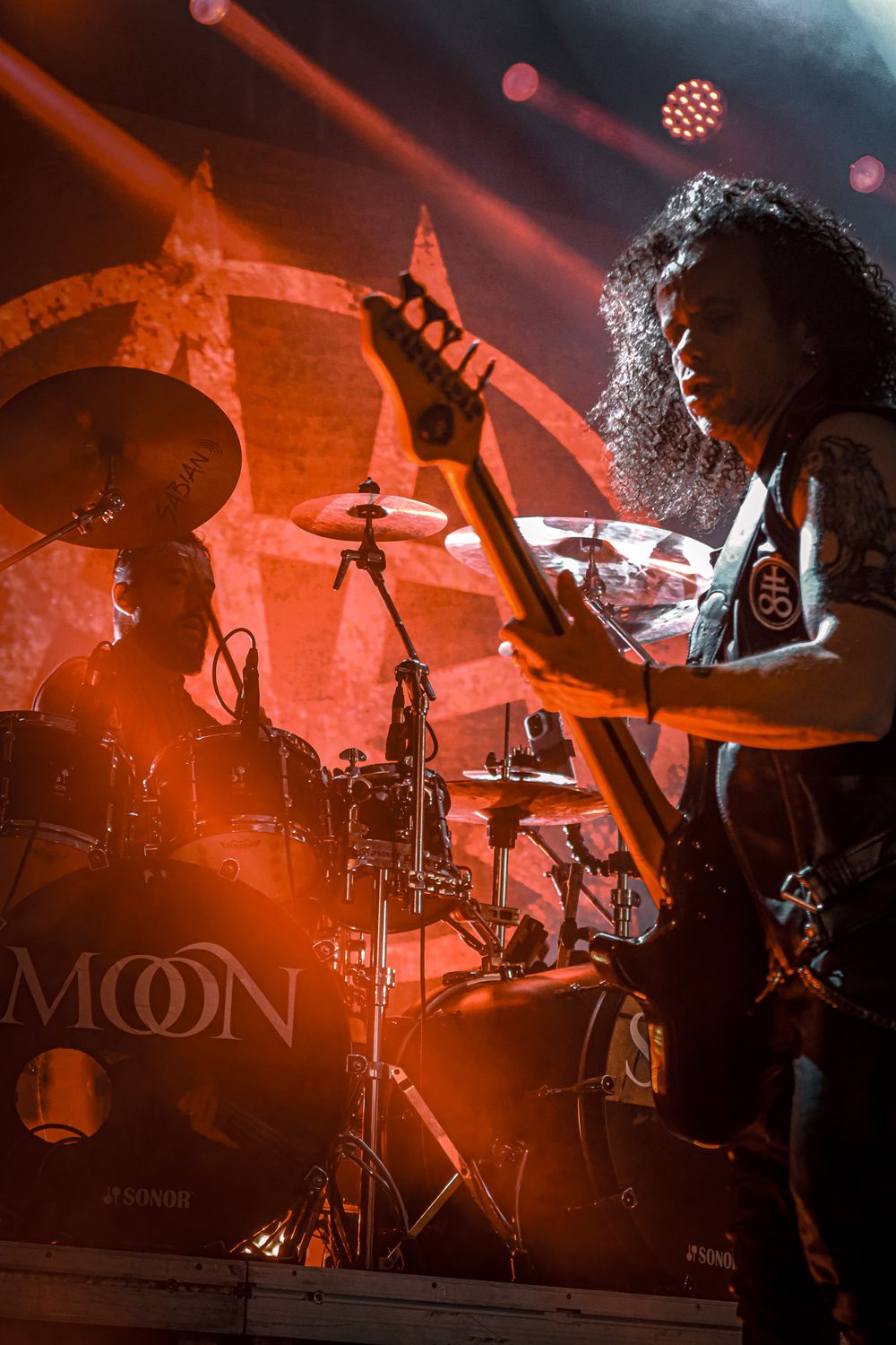 Drummer performs energetically behind drum kit with orange stage lighting and smoke effects.