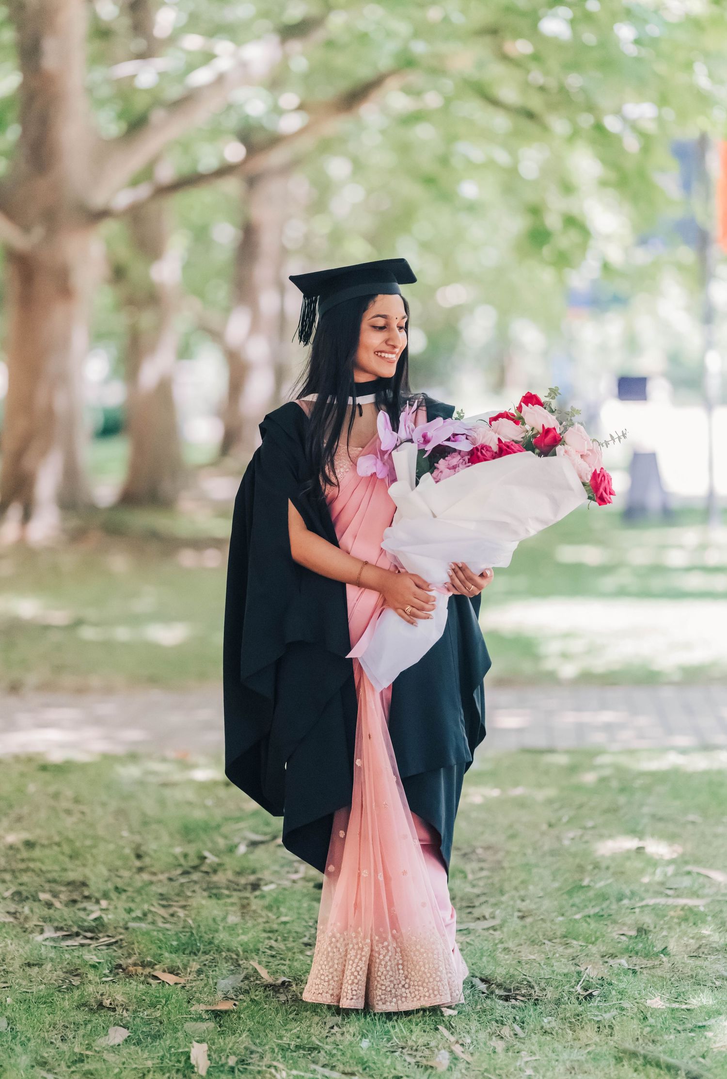 A graduate in black cap and gown holding a pink floral bouquet poses in a park setting with soft natural lighting.