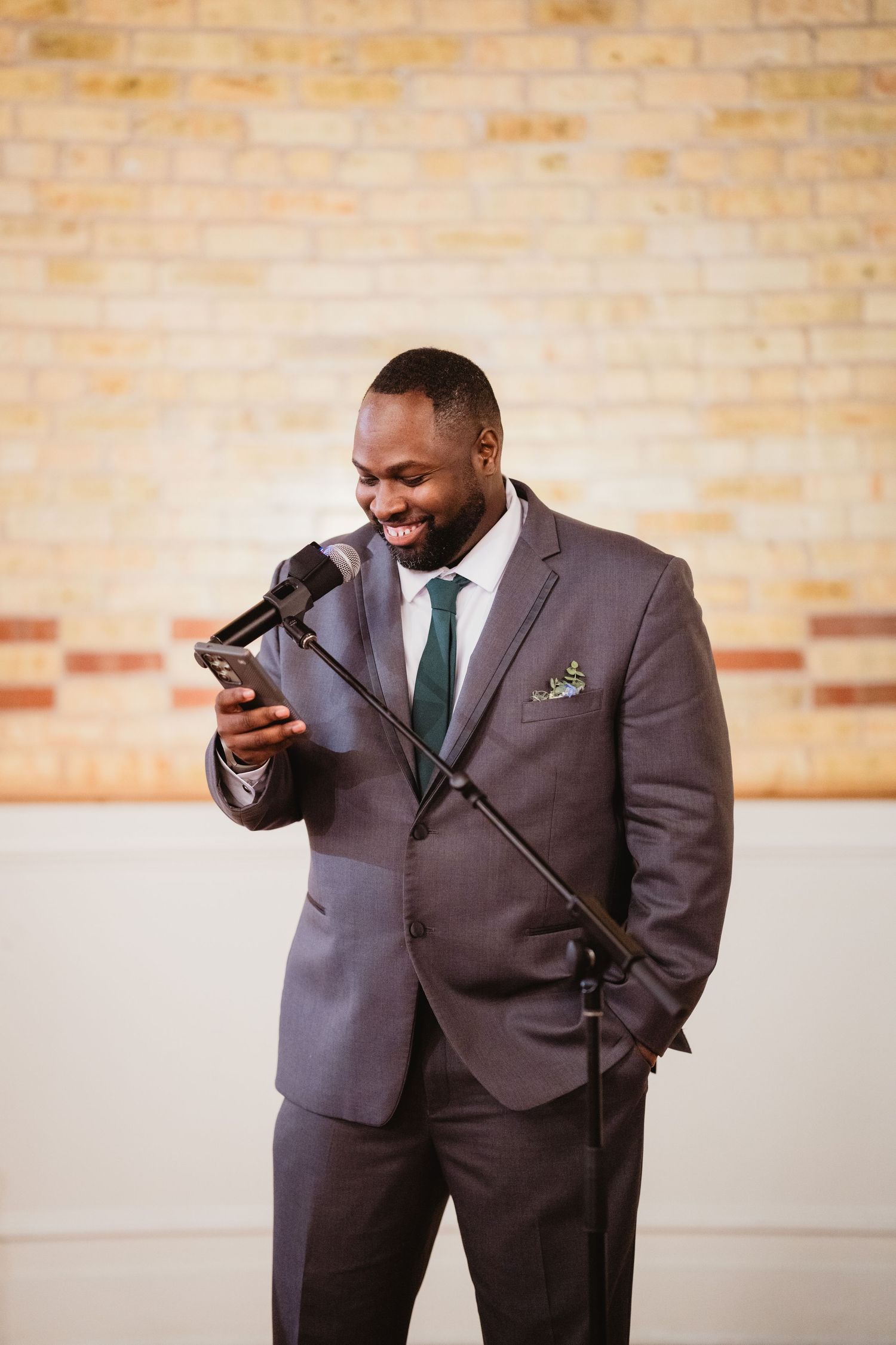 A person in a dark suit and green tie laughs while looking at their phone during what appears to be a wedding ceremony.