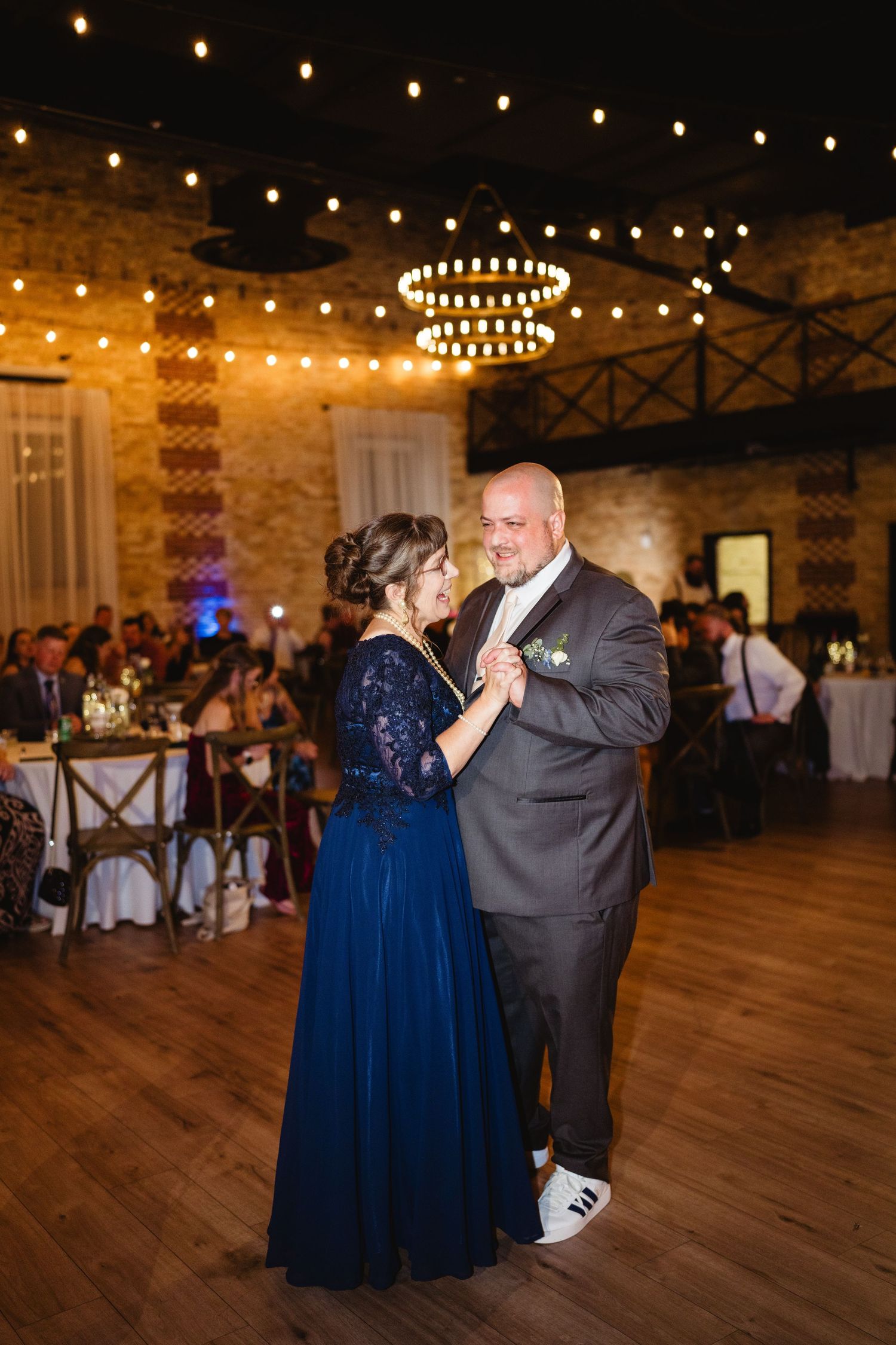 A couple shares a dance on a rustic wooden floor under string lights and a chandelier at a wedding reception.