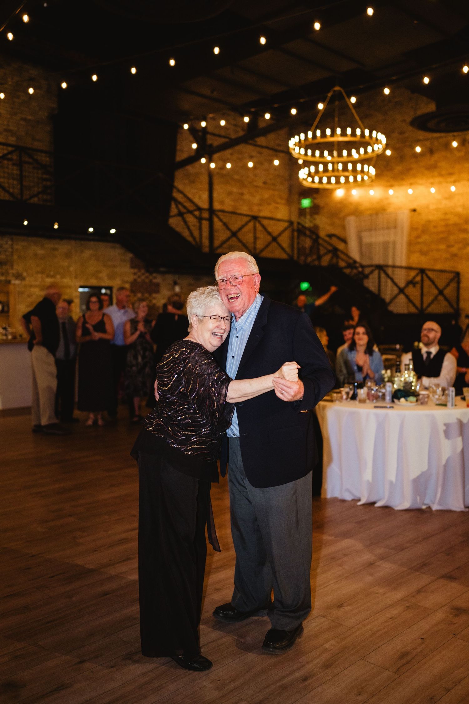 Couples slow dancing at an elegant wedding reception in a rustic venue with string lights overhead.