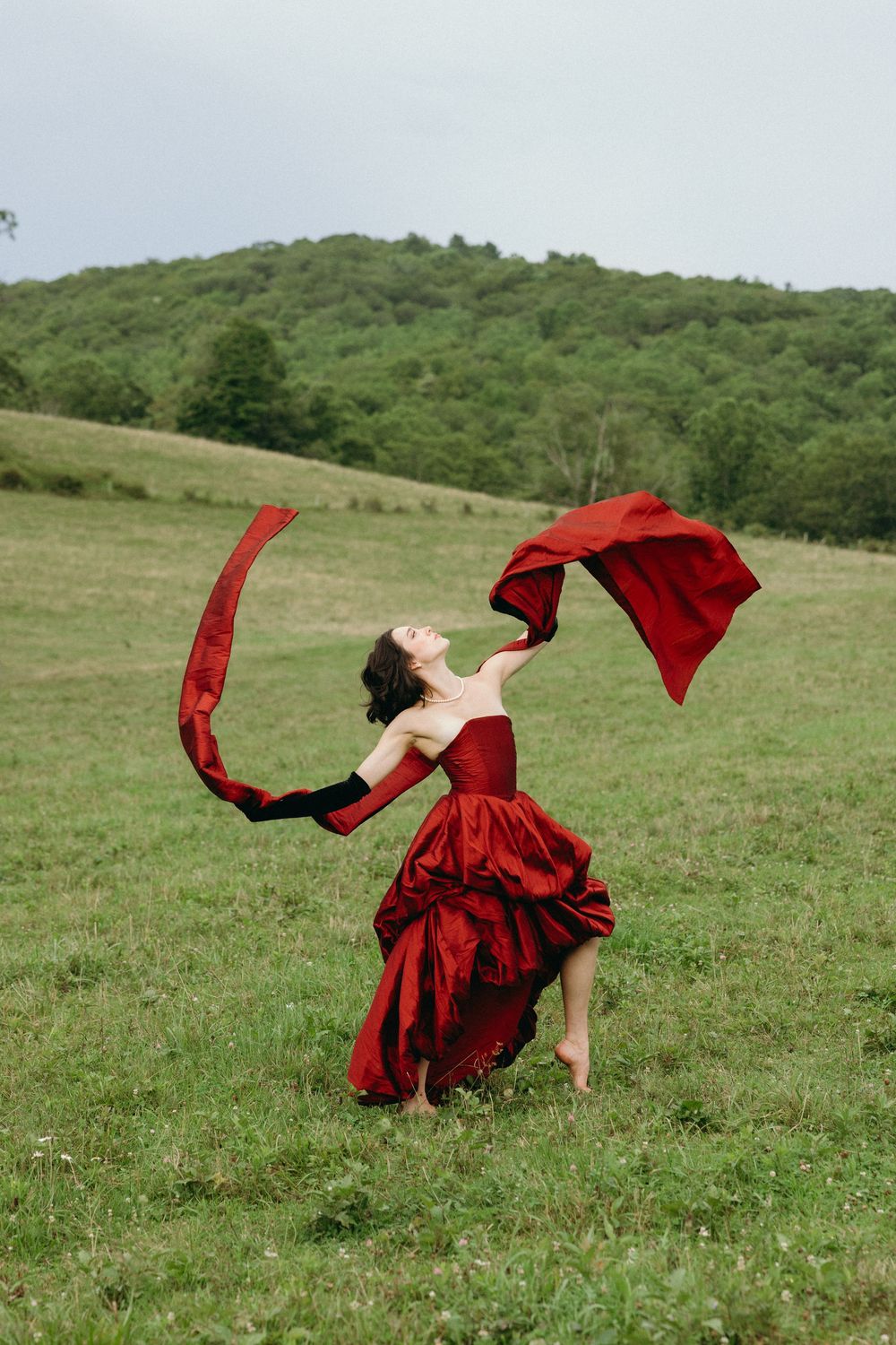 A woman in a flowing red dress dances and spins dramatically across a green hillside meadow in a sequence of movements.