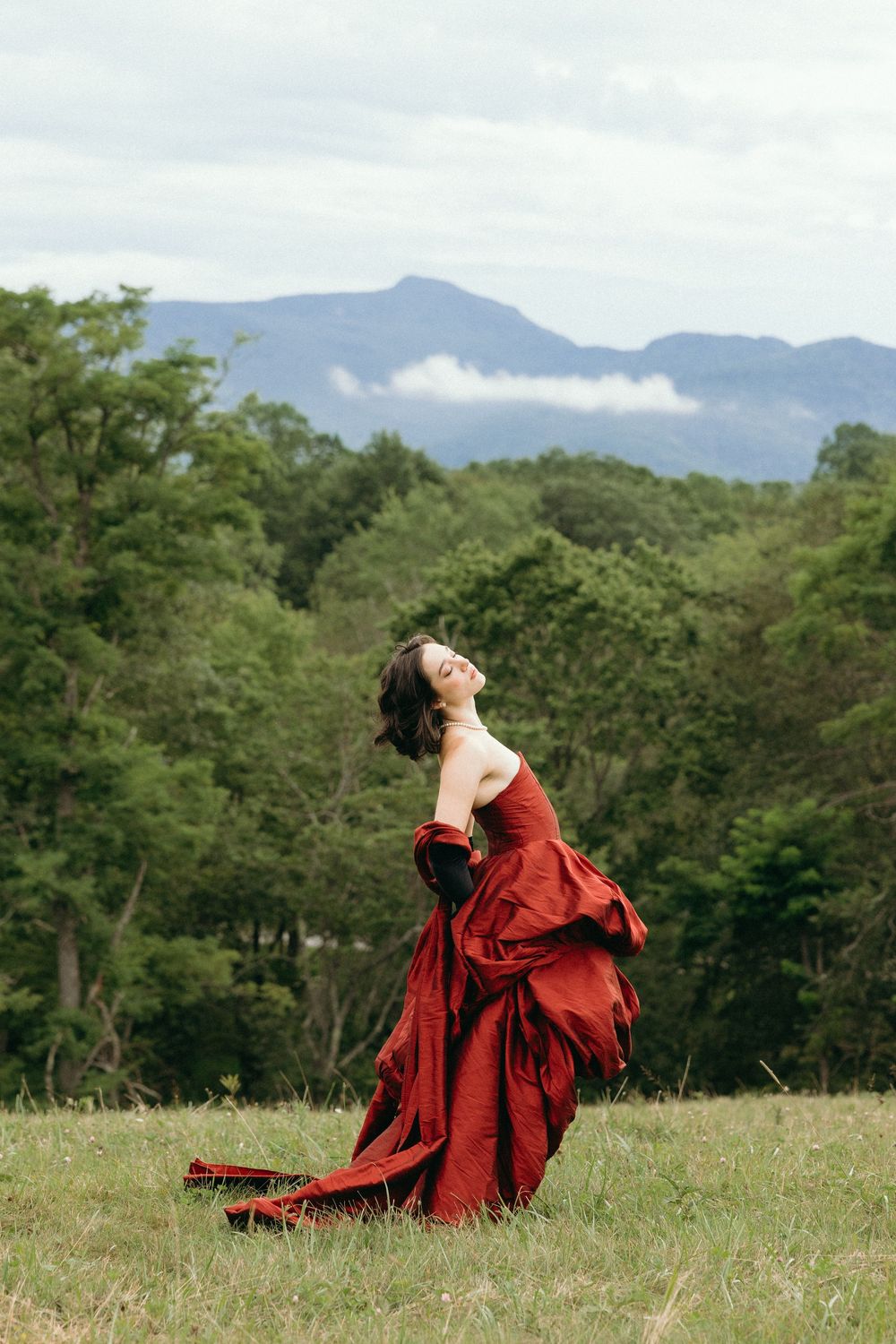 A woman in a flowing red dress sits gracefully in a meadow with mountains and trees creating a scenic backdrop.