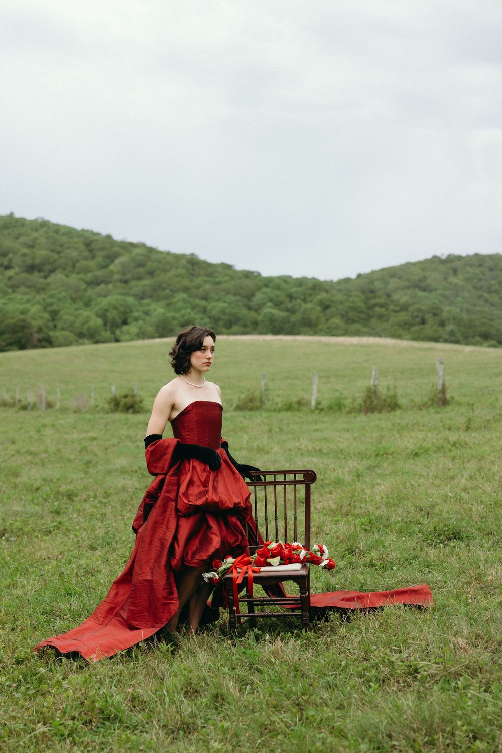 A woman in a flowing red gown sits beside an antique chair in a serene meadow with green hills in the background.