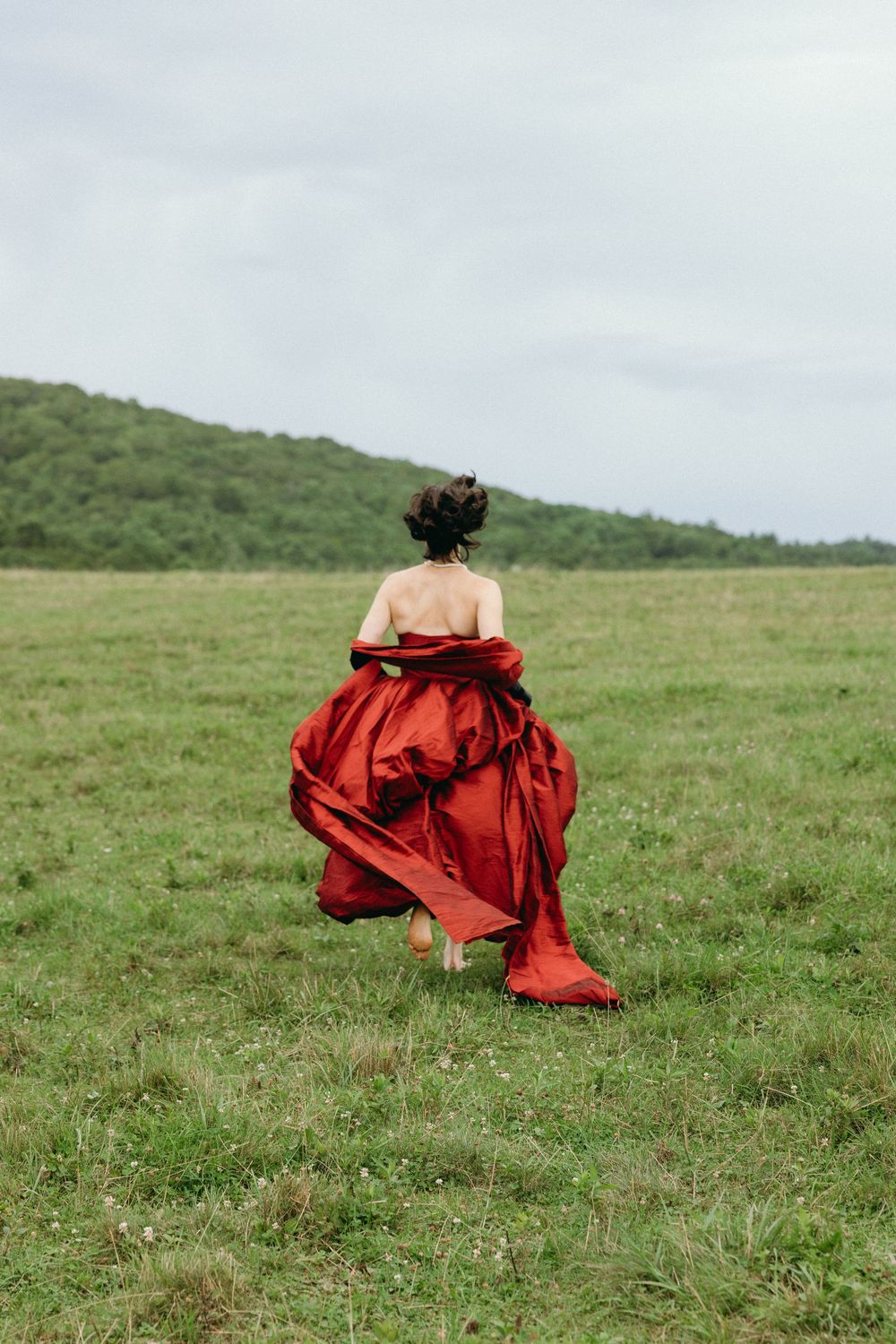 a woman in a flowing red dress and boots walks across a green meadow with rolling hills in the background.