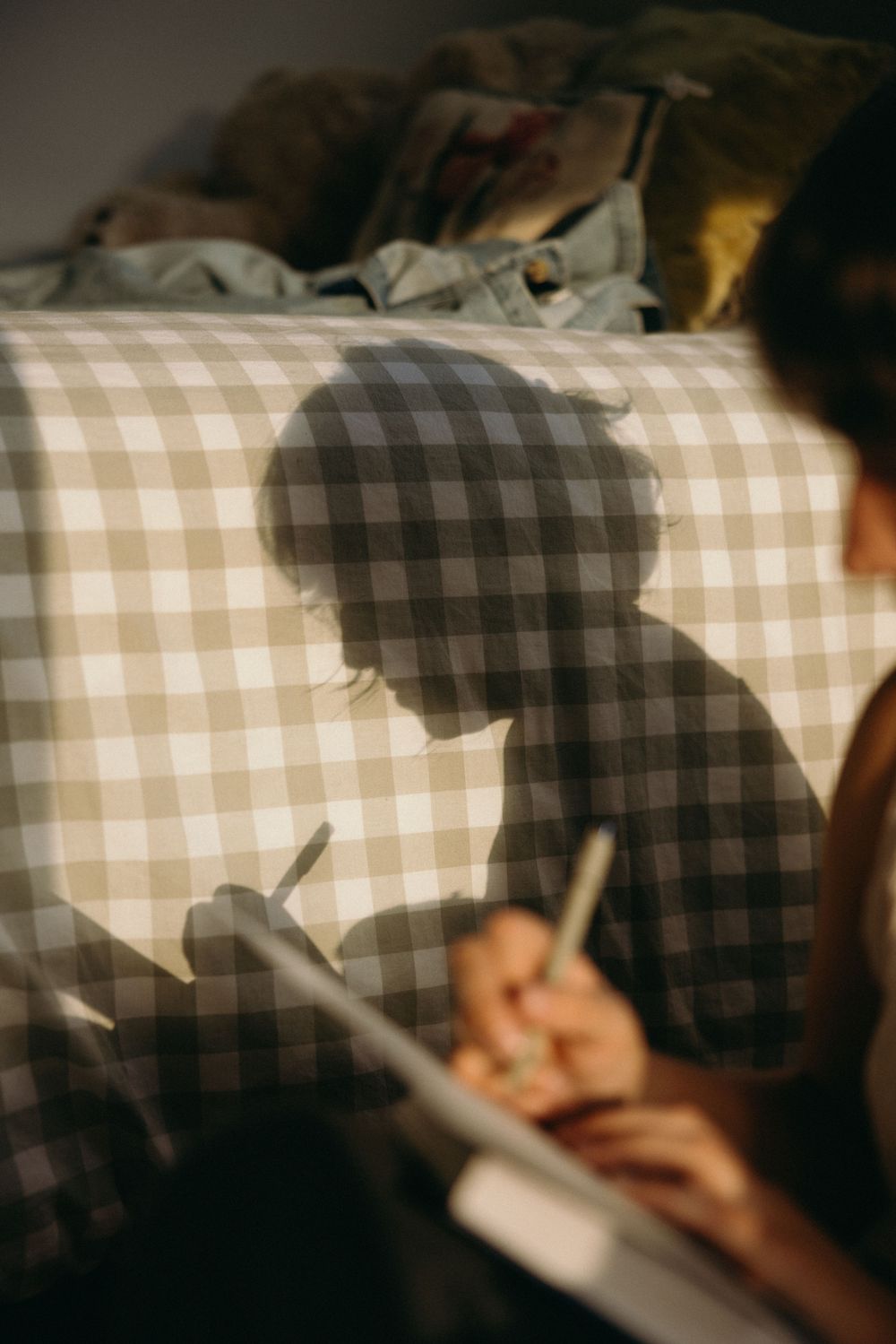 Person wearing a plaid shirt writes in a notebook while sitting at a desk in dim lighting.