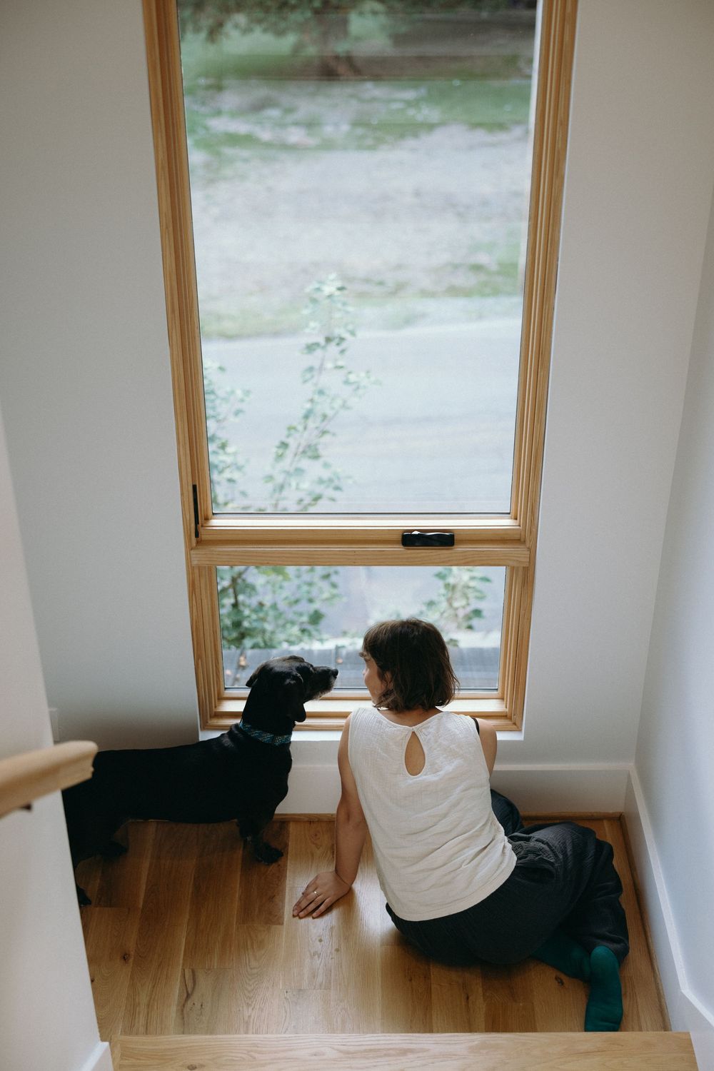 Person in white shirt sits by window in minimalist room with wooden floor and white walls.
