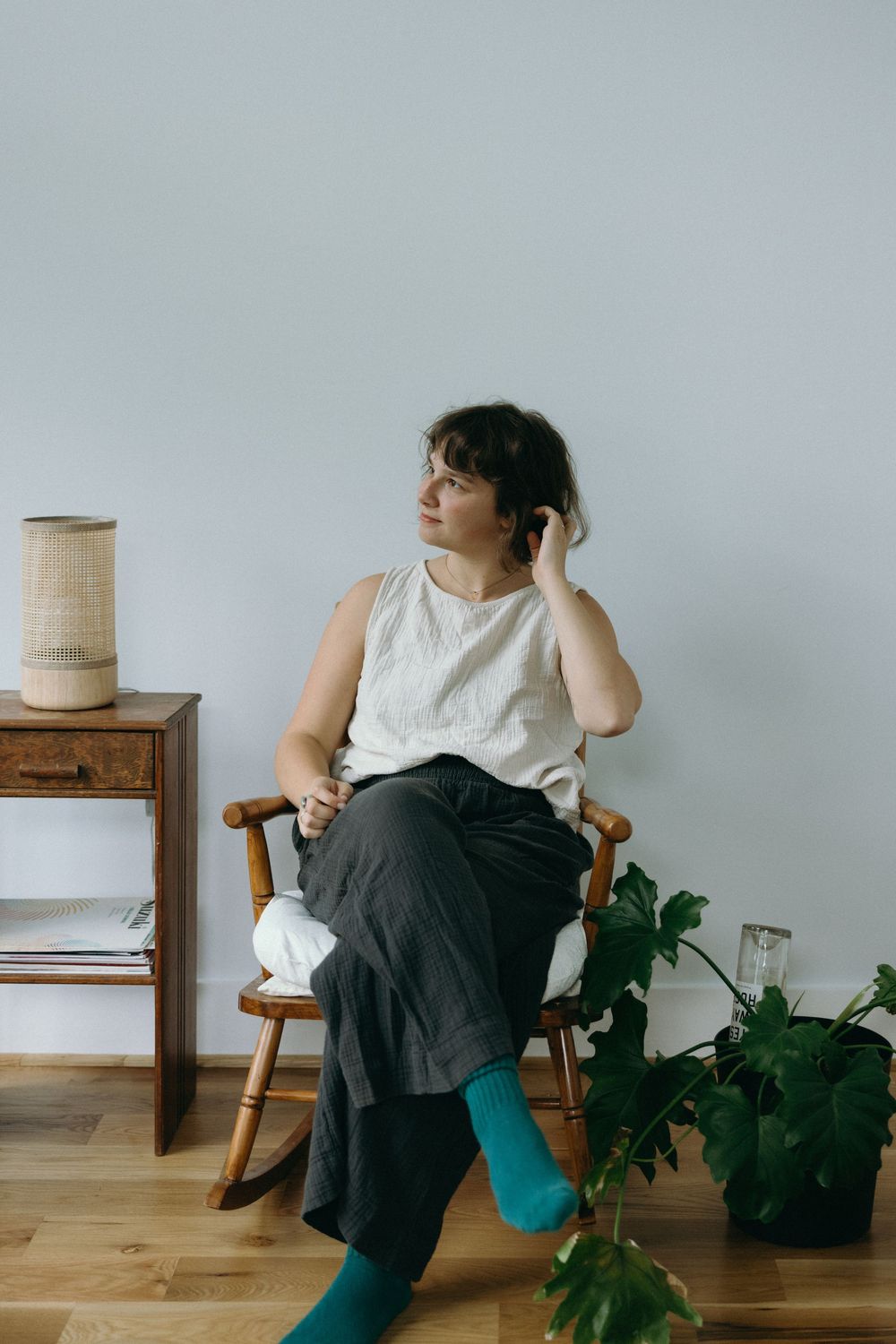 A person in a white top and black skirt sits thoughtfully in a wooden chair beside houseplants against a light gray wall.