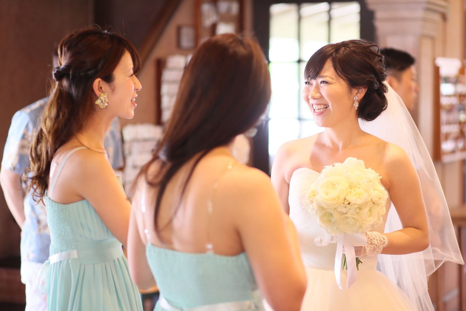 Smiling bride in white gown holding bouquet chats with bridesmaids in mint dresses at wedding ceremony.
ブライズメイド