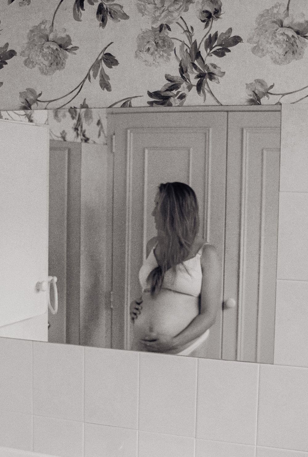 Artistic black and white photograph showing floral wallpaper pattern and bathroom fixtures in soft lighting.