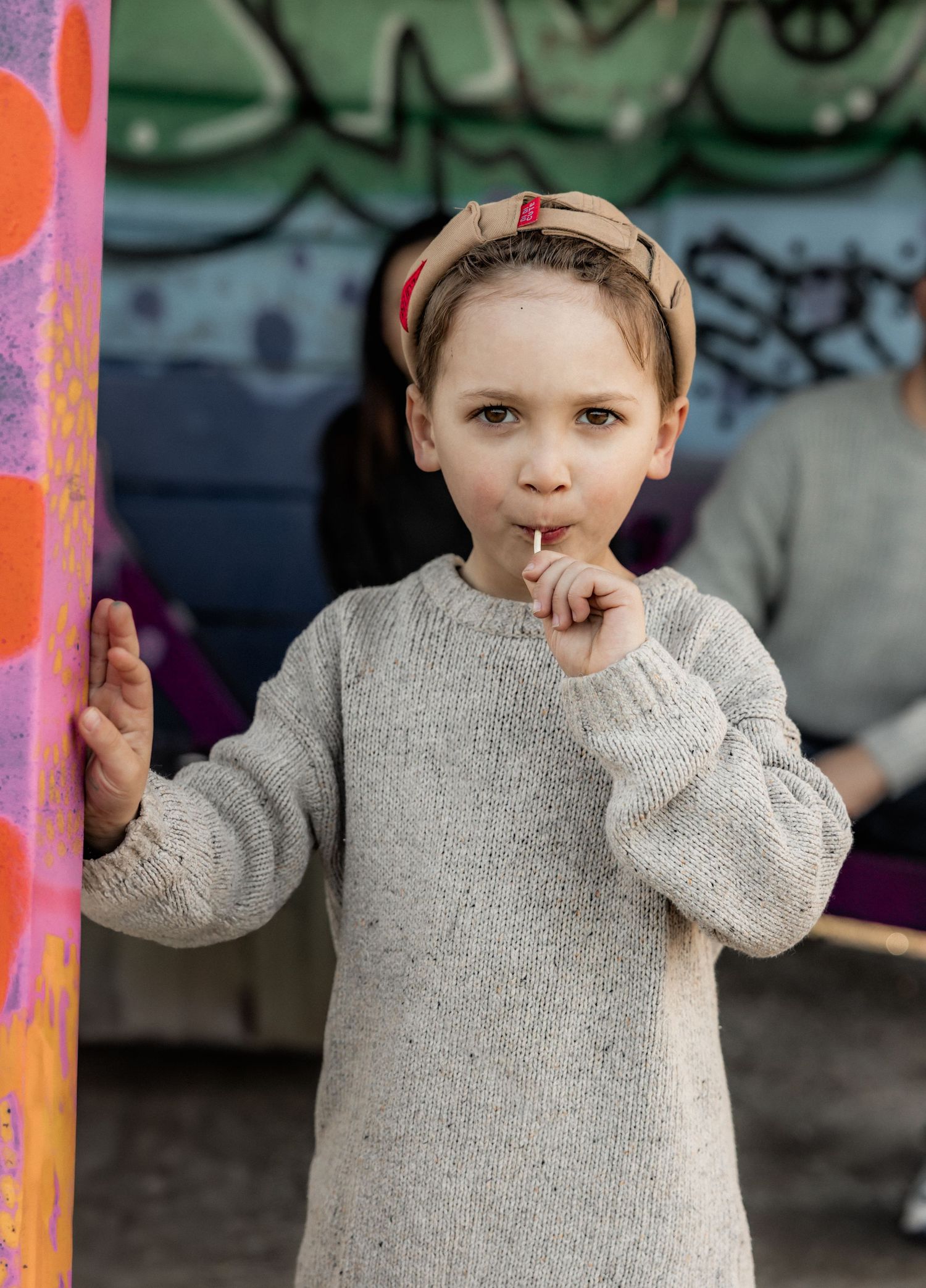 Child in grey sweater posing against colorful graffiti wall.