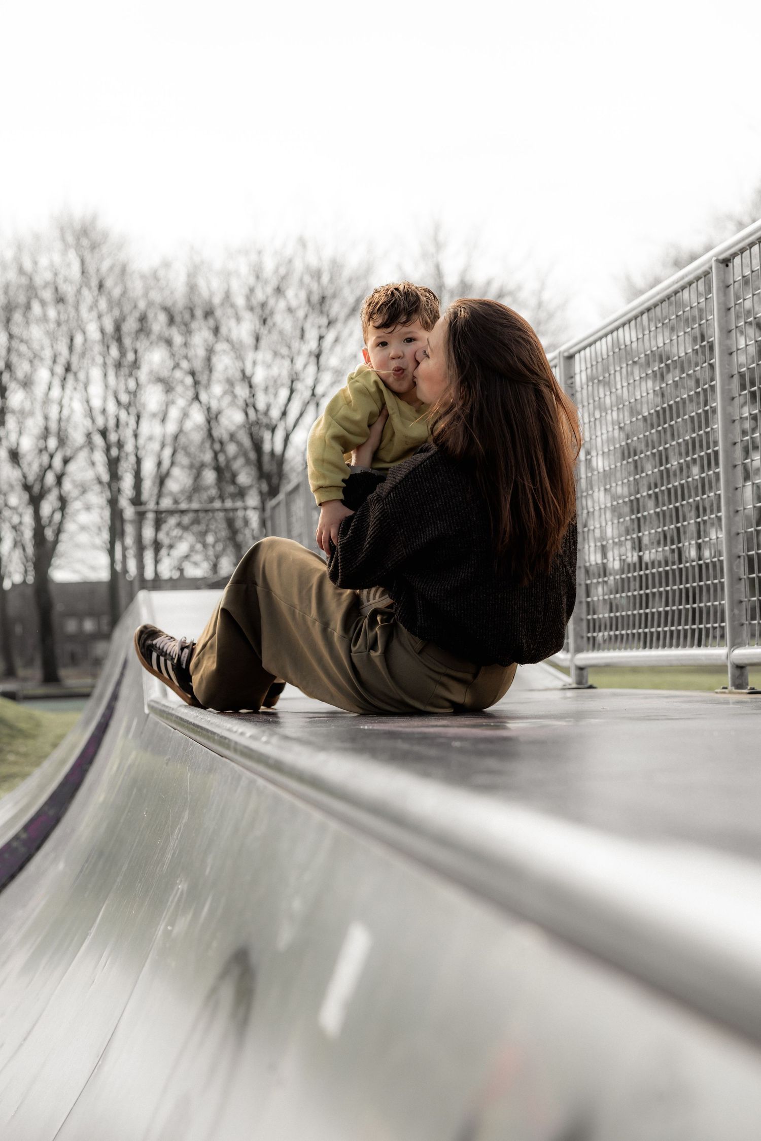 People sliding down a curved metal ramp at a skate park on an overcast day.