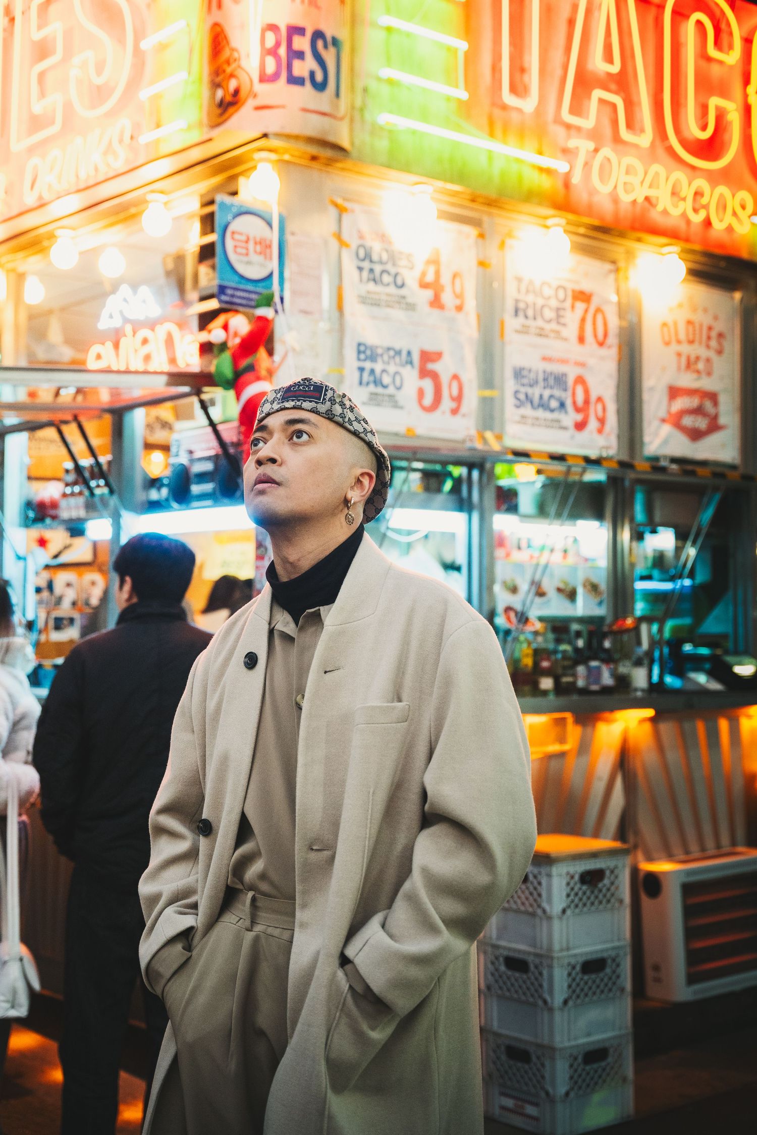 Three people in beige coats stand near colorful neon signs at a night market.