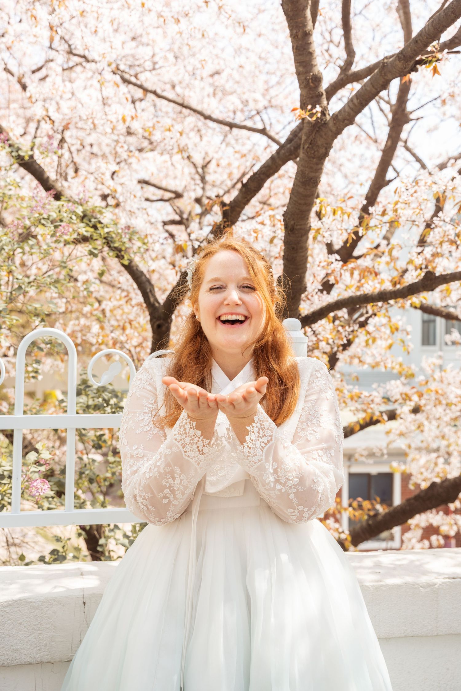 A joyful person in a white lace dress poses under blooming cherry blossom trees in springtime.