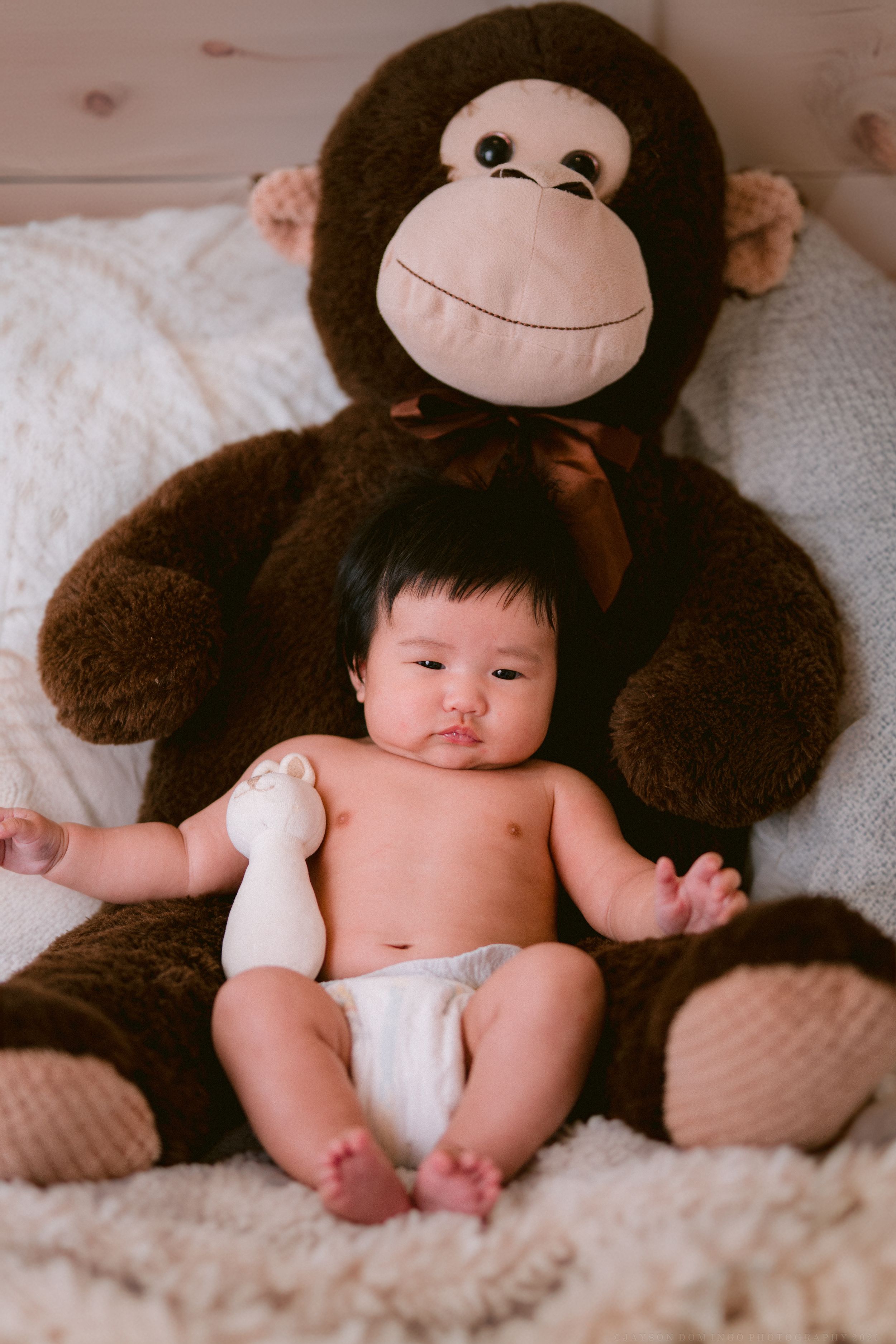Baby rests on plush white bedding next to a large brown stuffed monkey toy.