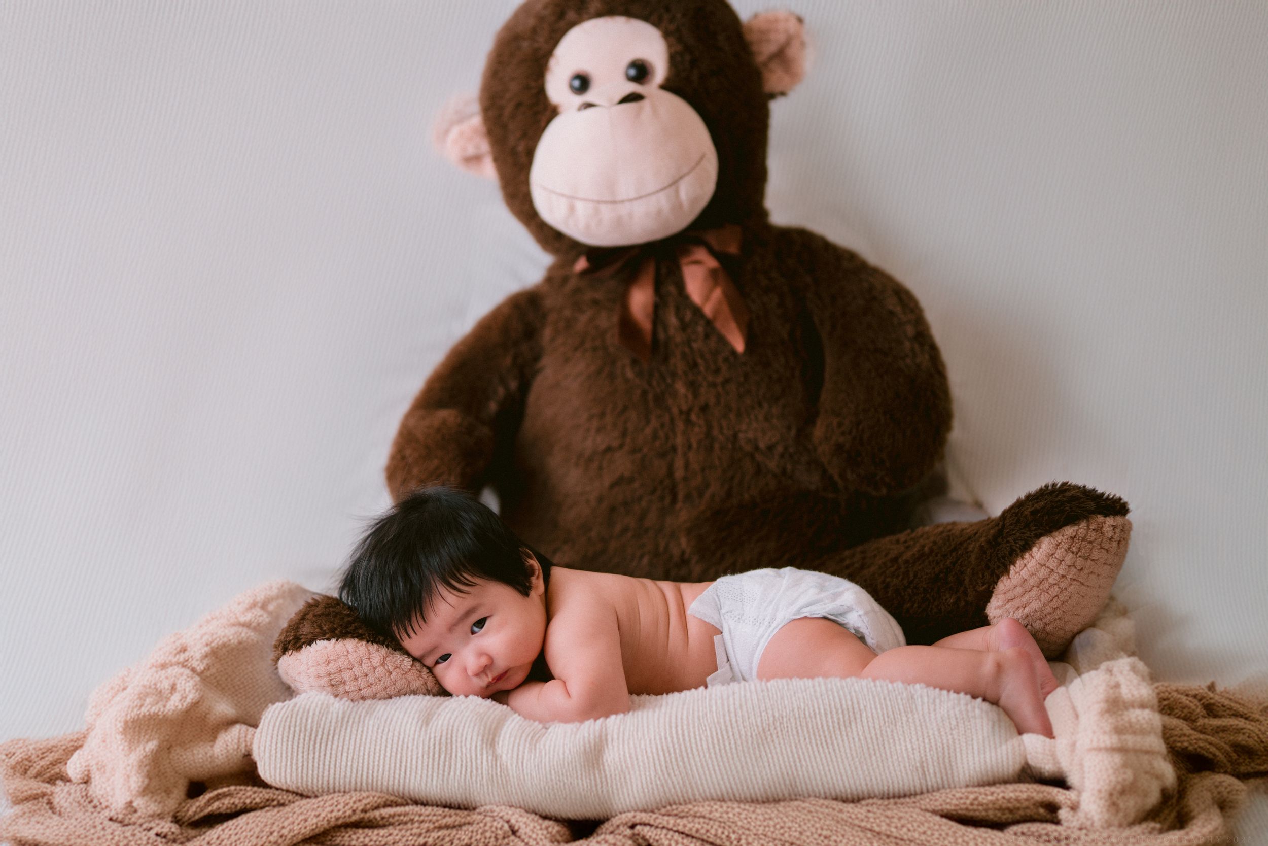 Baby resting beside large brown plush monkey on white bedding.