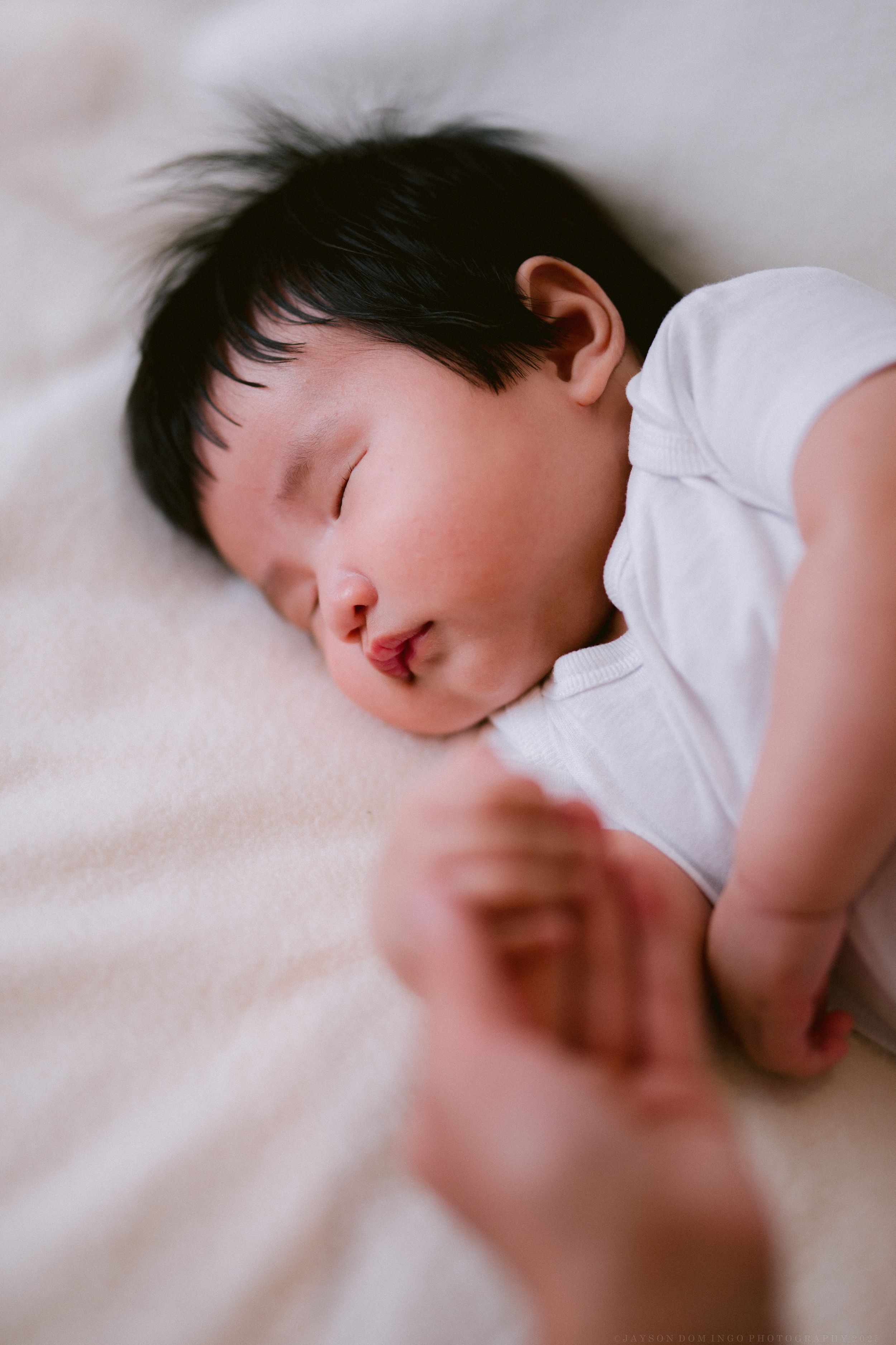 Sleeping baby with dark hair rests peacefully on white bedding in soft natural light.