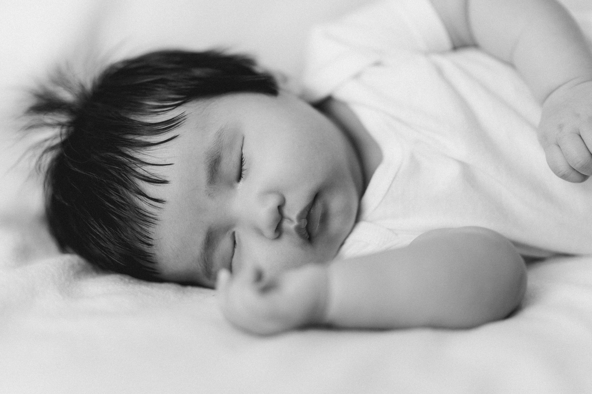 Sleeping baby in white clothing rests peacefully on light bedding.