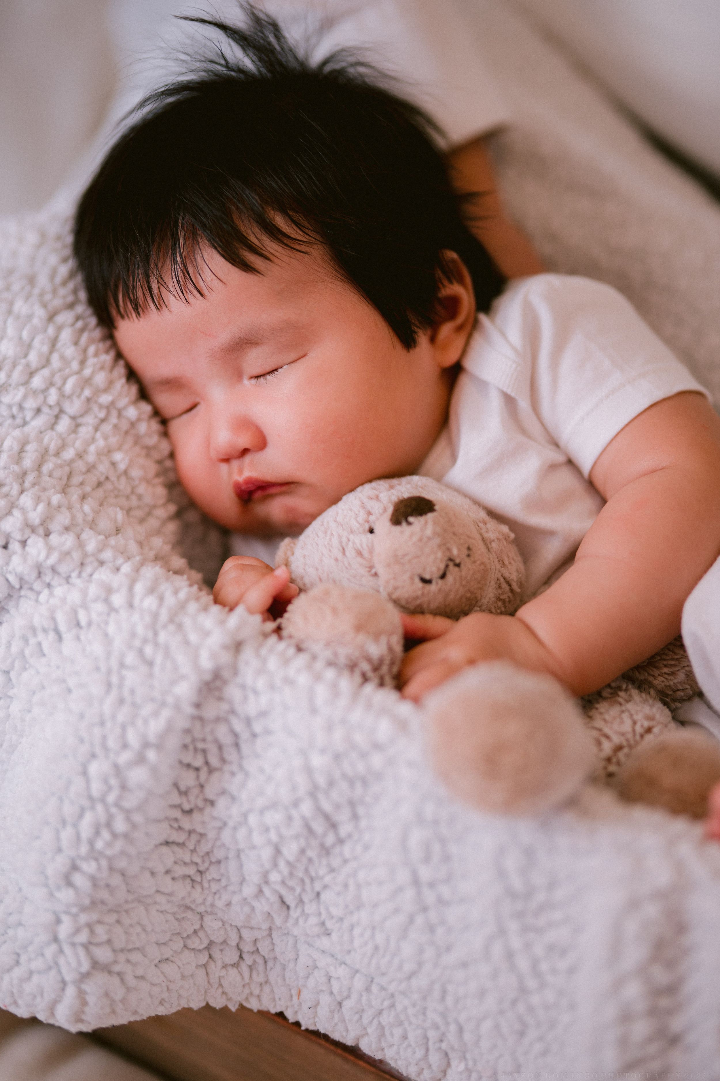 A baby in white sleeps soundly while cuddling with a plush teddy bear on fluffy white bedding.