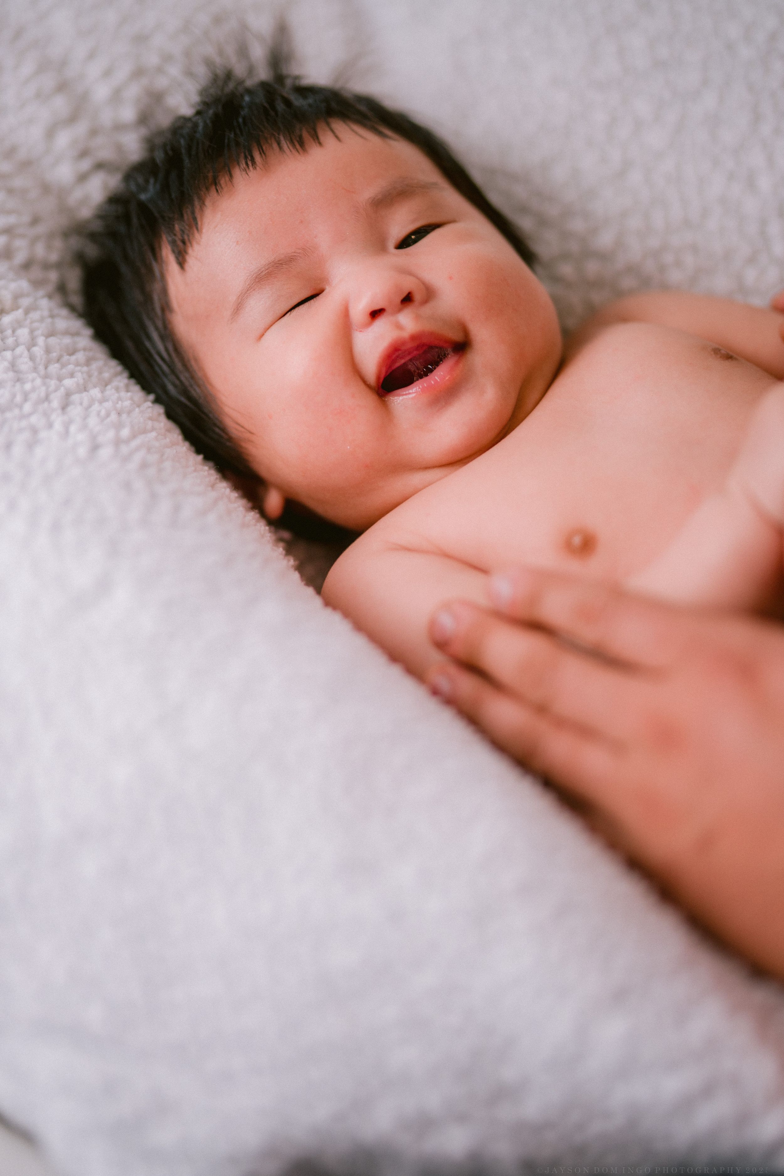 A joyful baby lies on soft white bedding while laughing with bright eyes closed.