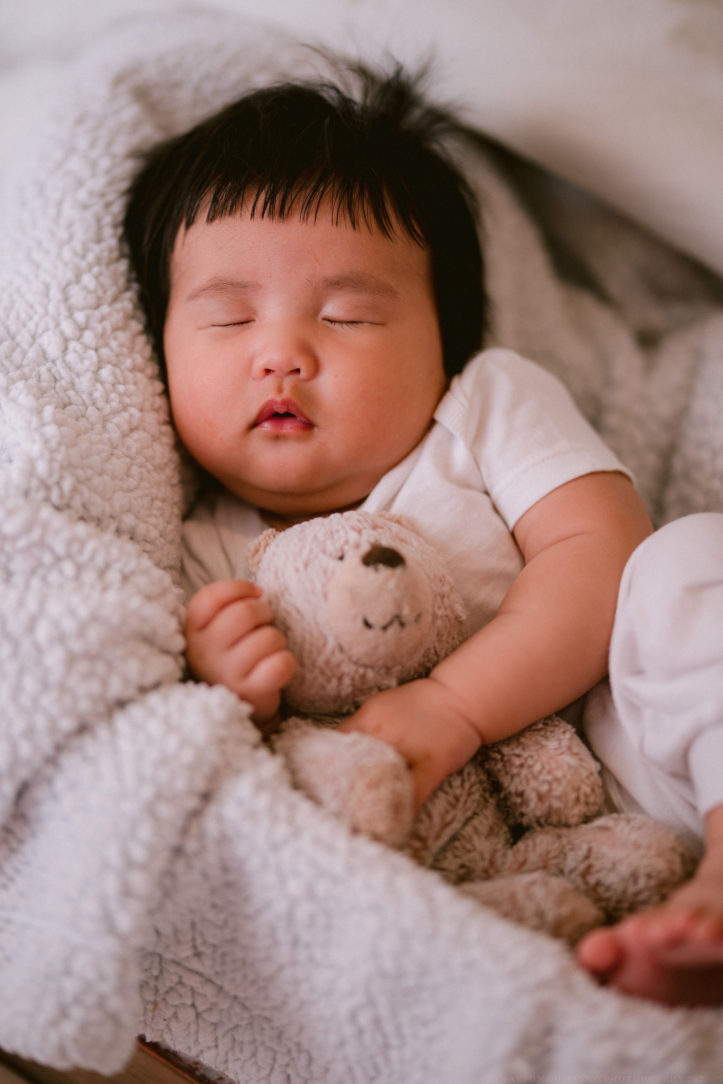 Baby sleeping peacefully with plush toy under soft white blanket.