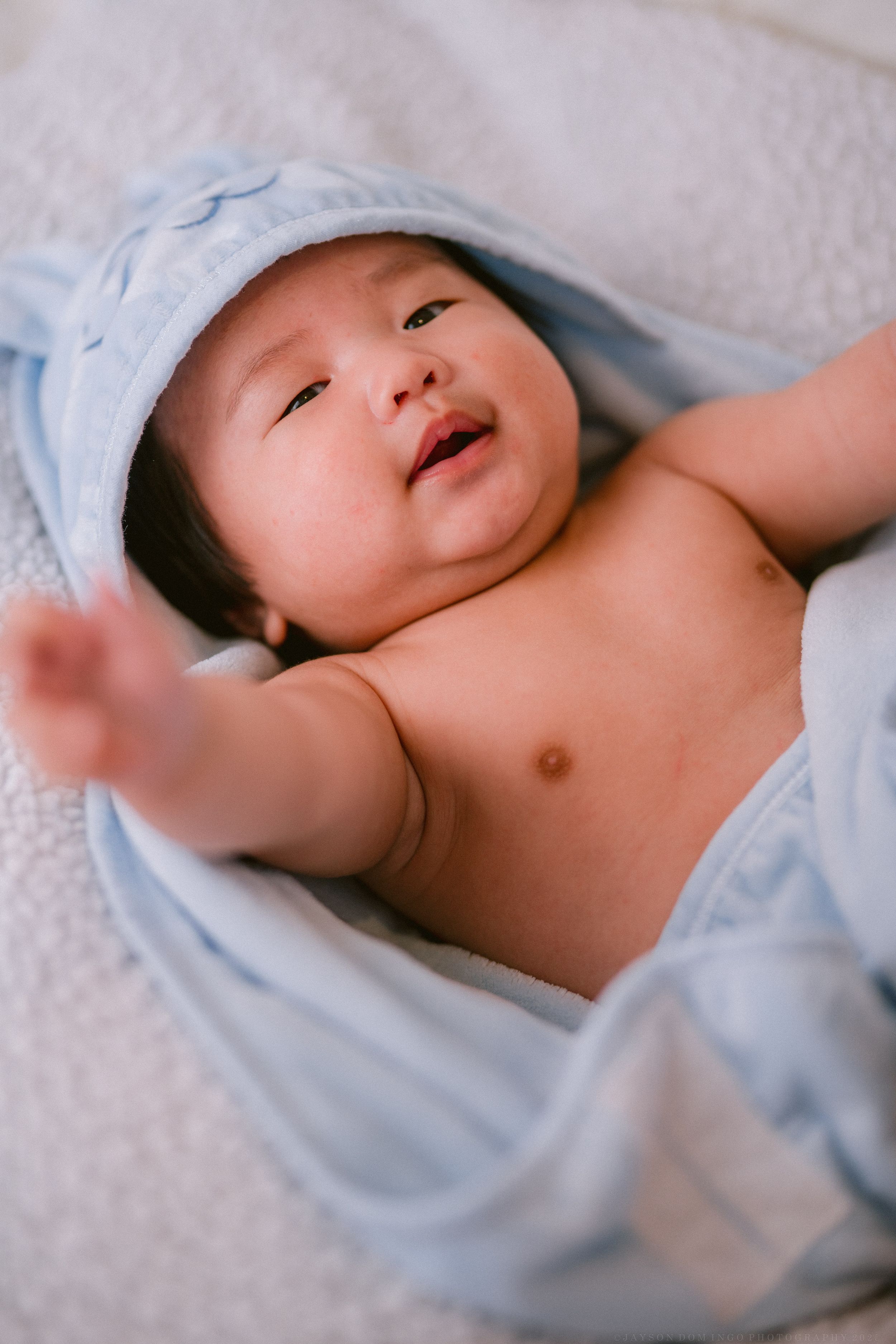 Baby wearing light blue clothing lies on white bedding while smiling and stretching.