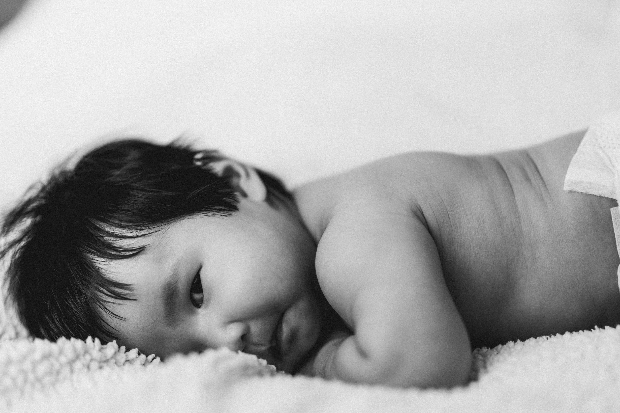 Infant lying on plush white bedding in gentle monochrome lighting.