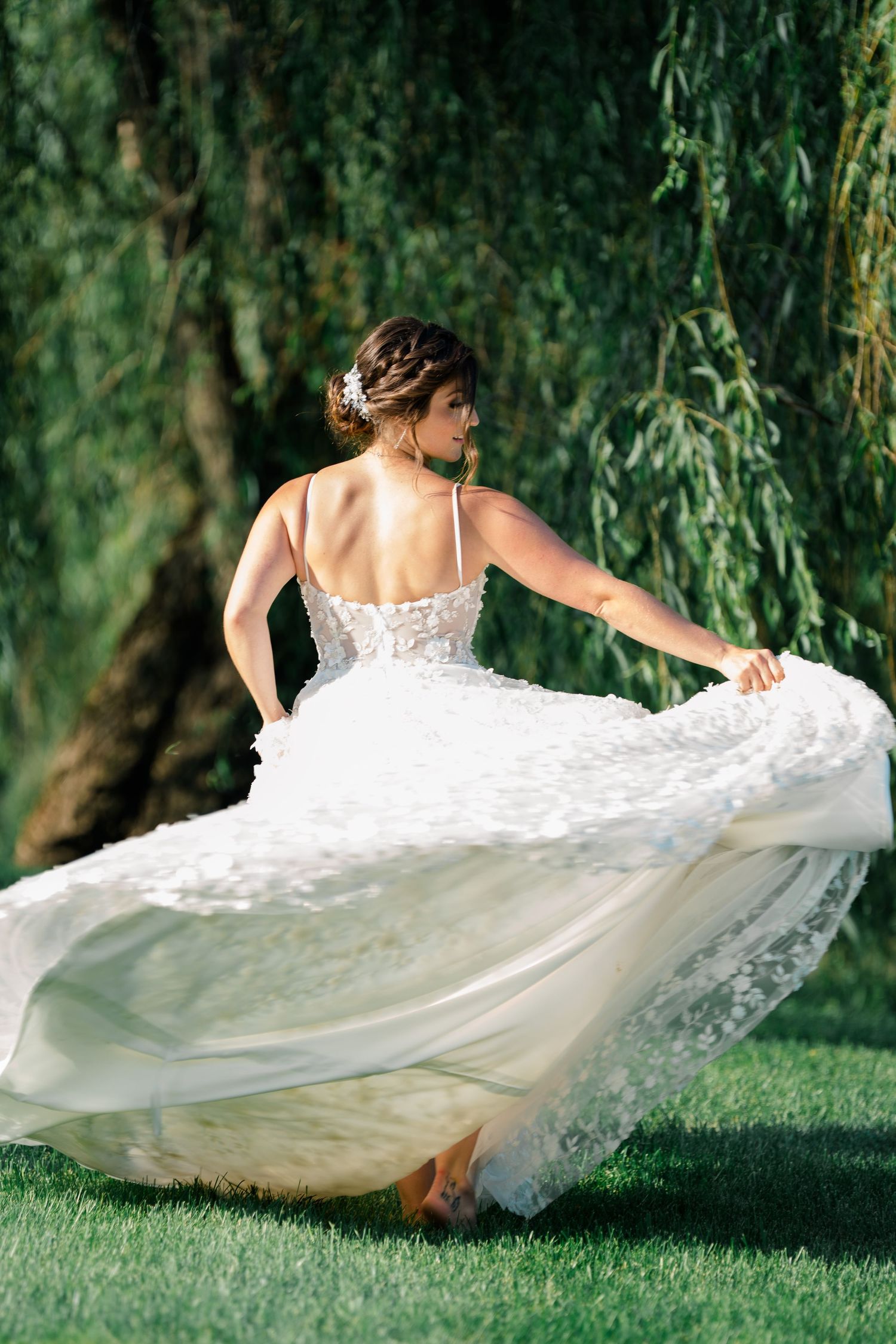 Bride in flowing white lace dress twirling on green lawn beneath willow trees.
