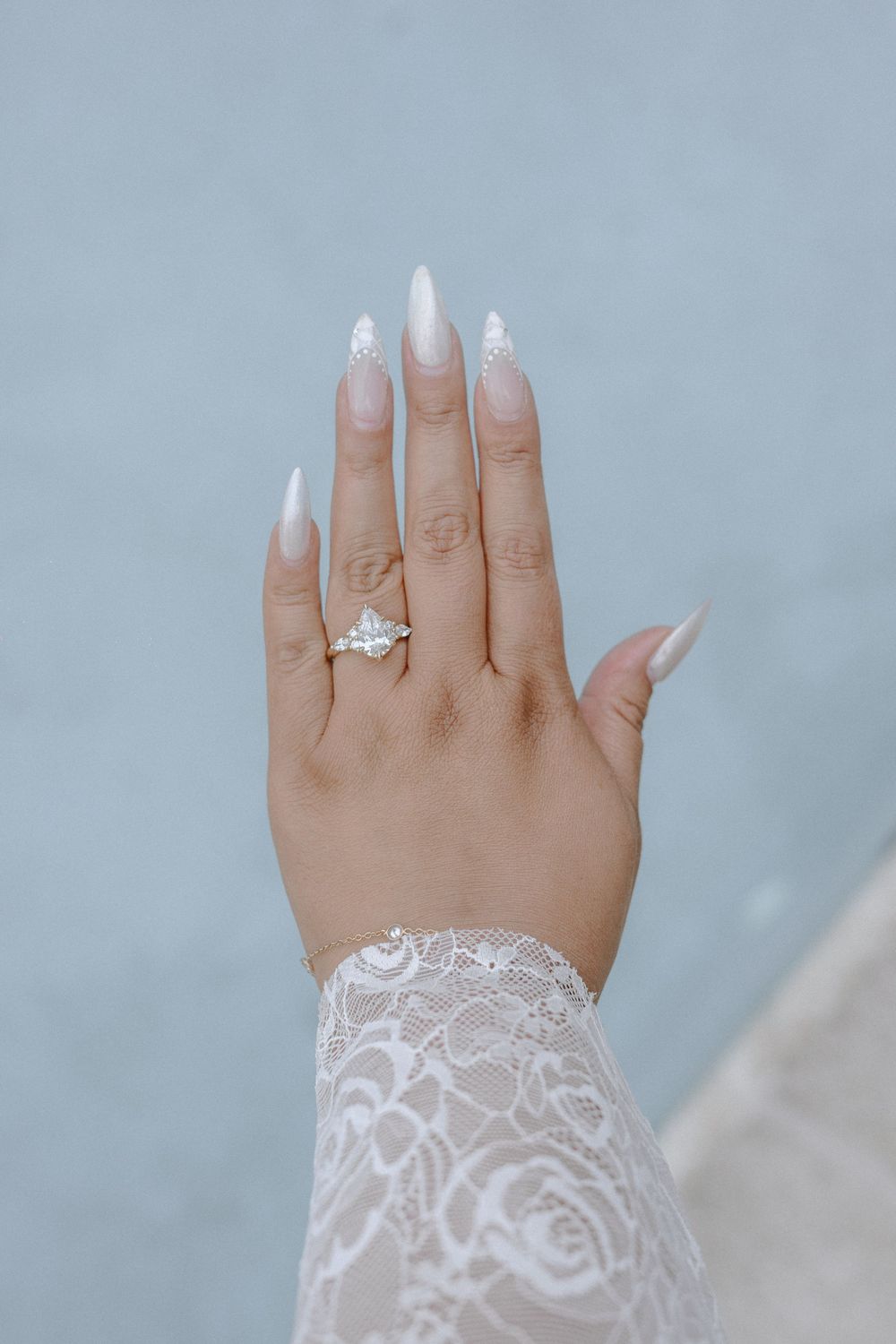 Hand wearing delicate lace sleeve displays elegant manicured nails and sparkling engagement ring against soft gray background.