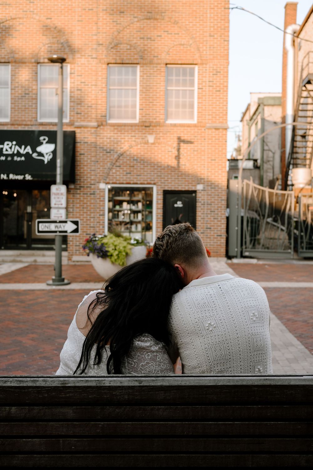 A couple sits close together on a bench outside a brick building on a cobblestone street at sunset.