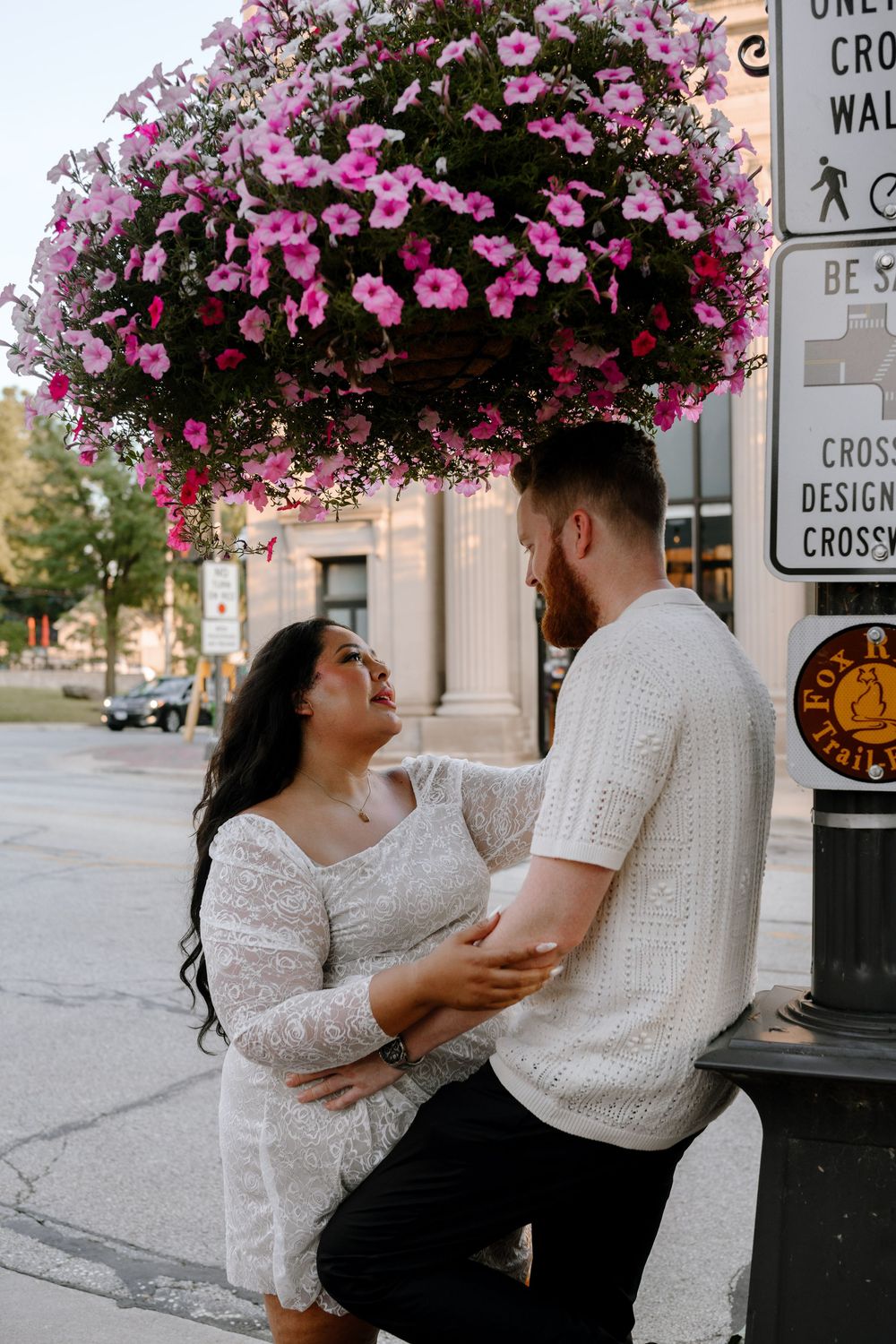 A couple shares romantic moments beneath vibrant pink floral hanging baskets on a city street.