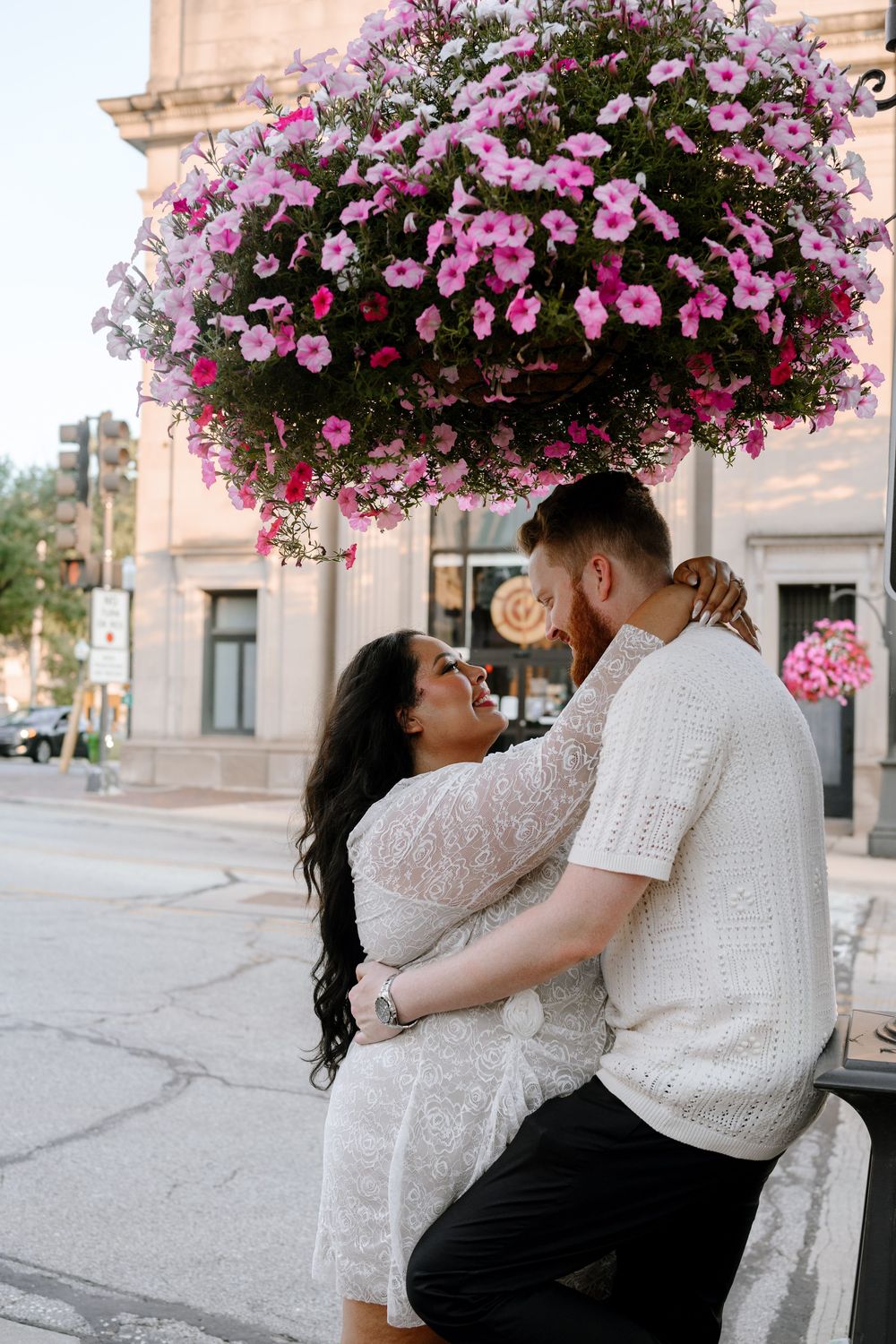 A couple embraces under beautiful hanging pink petunias on a downtown street corner during a romantic moment.