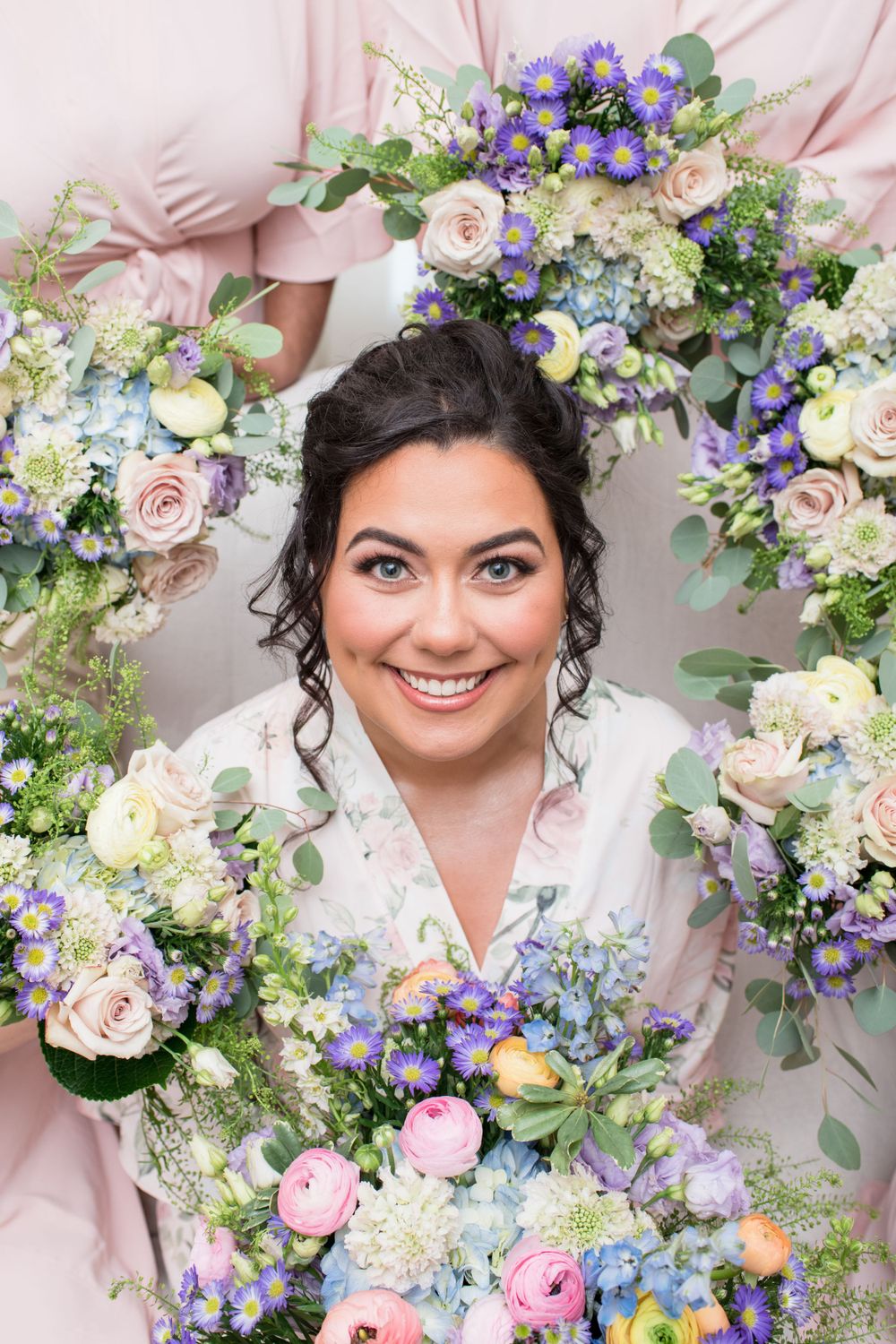 Beautiful floral arrangement with pink roses, blue delphinium, and white hydrangeas surrounds a bridal portrait in soft lighting.