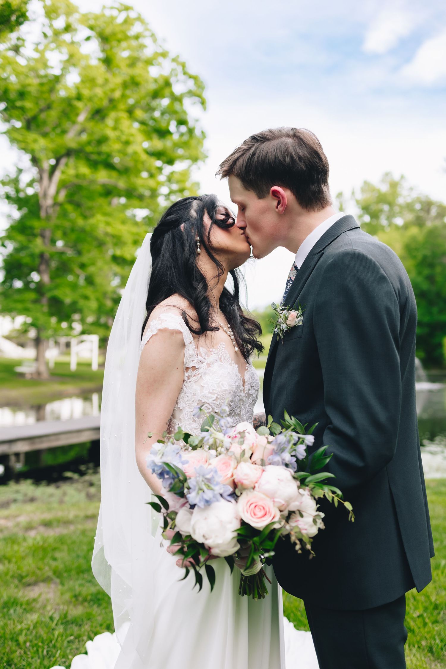 Newlyweds share a romantic kiss in an outdoor garden setting with a beautiful bridal bouquet of pink and white flowers.