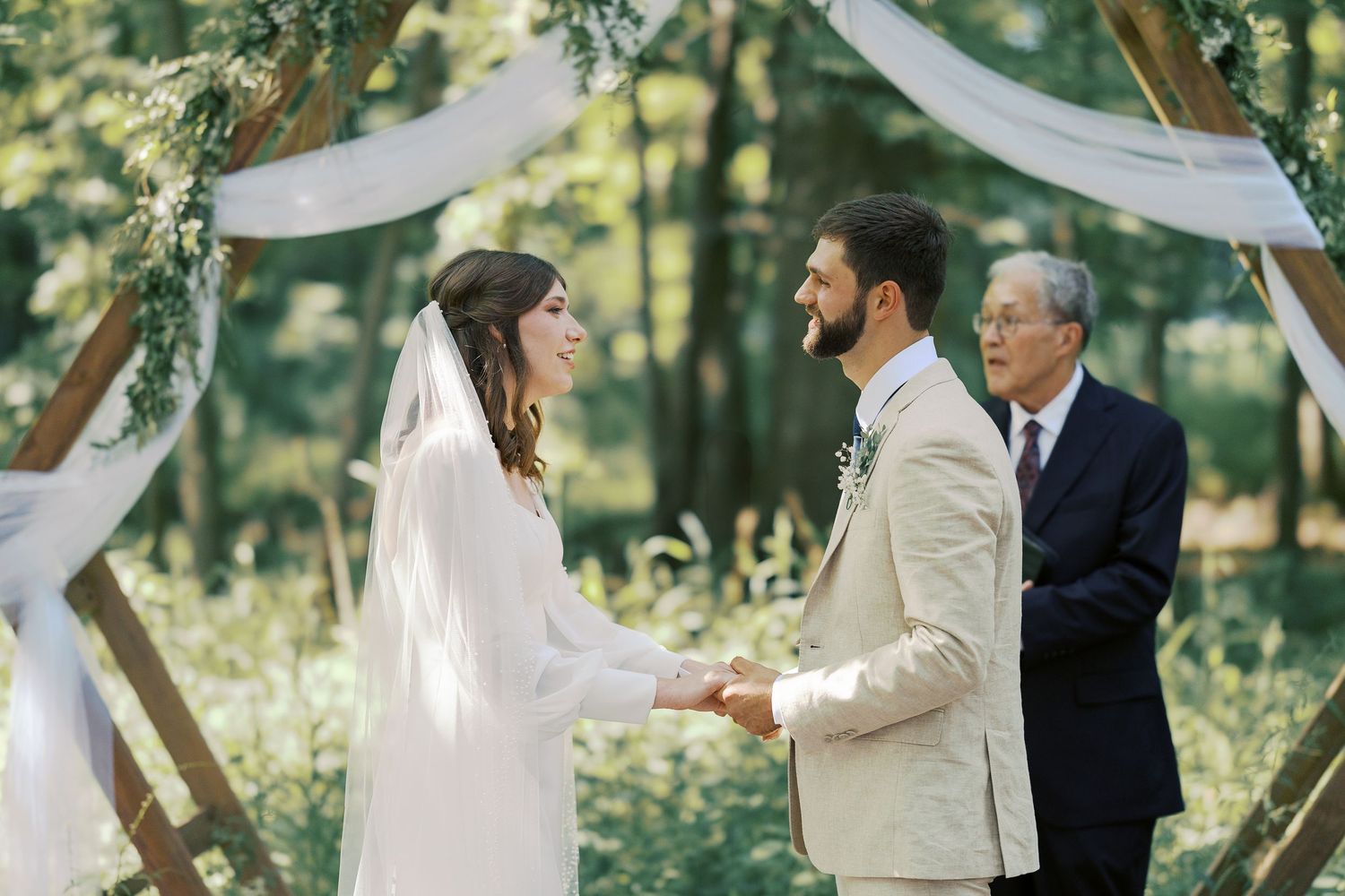 Wedding ceremony takes place under white draped fabric in a sunlit forest setting.