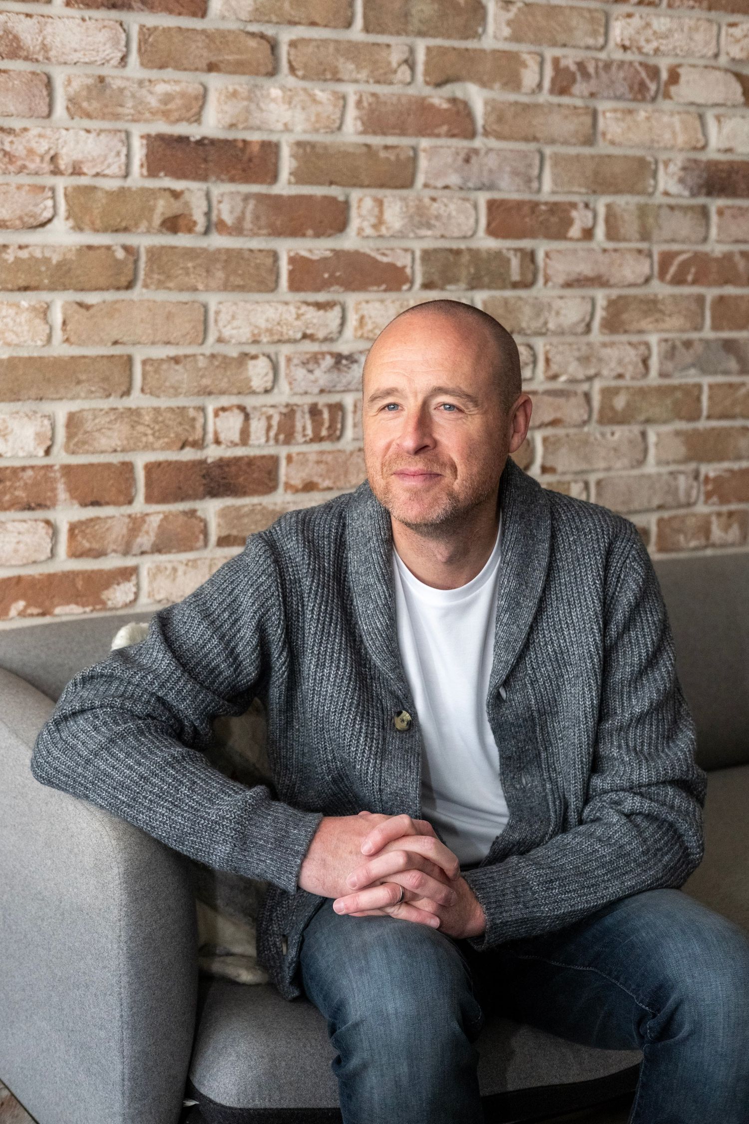 Someone wearing a gray cardigan sweater and white t-shirt sits casually in a gray armchair against an exposed brick wall.